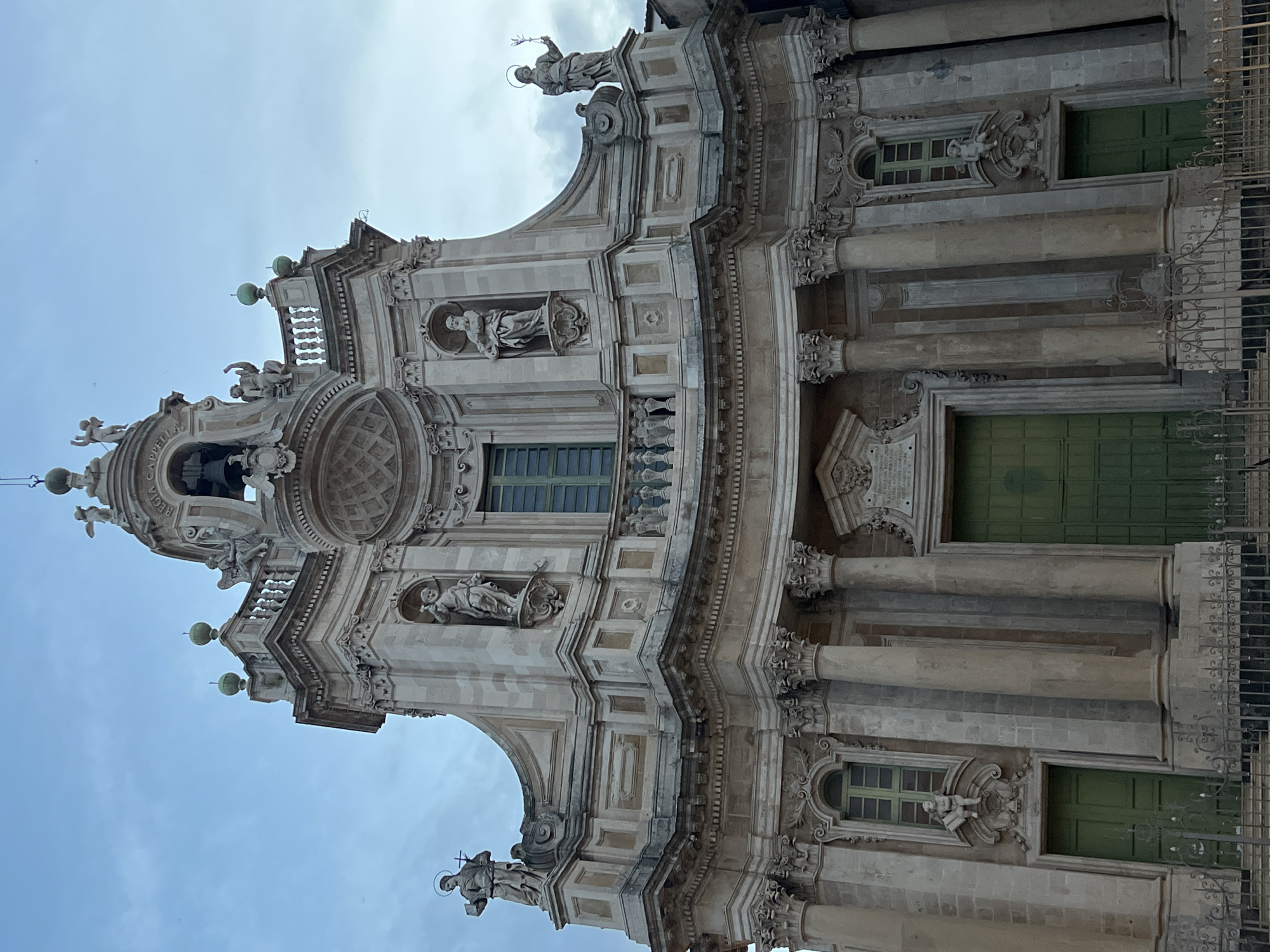 This is a photograph of the facade of the Church of Santa Chiara in Catania, Italy. The church is constructed of stone and features a Baroque architectural style. The facade is adorned with statues of saints and angels, ornate carvings, and decorative columns. At the top of the facade, there is a bell tower with a bell. The entrance to the church is through a large green double door. There are also two smaller green doors on either side of the main entrance. The church is surrounded by a decorative metal fence. The sky in the background is cloudy.