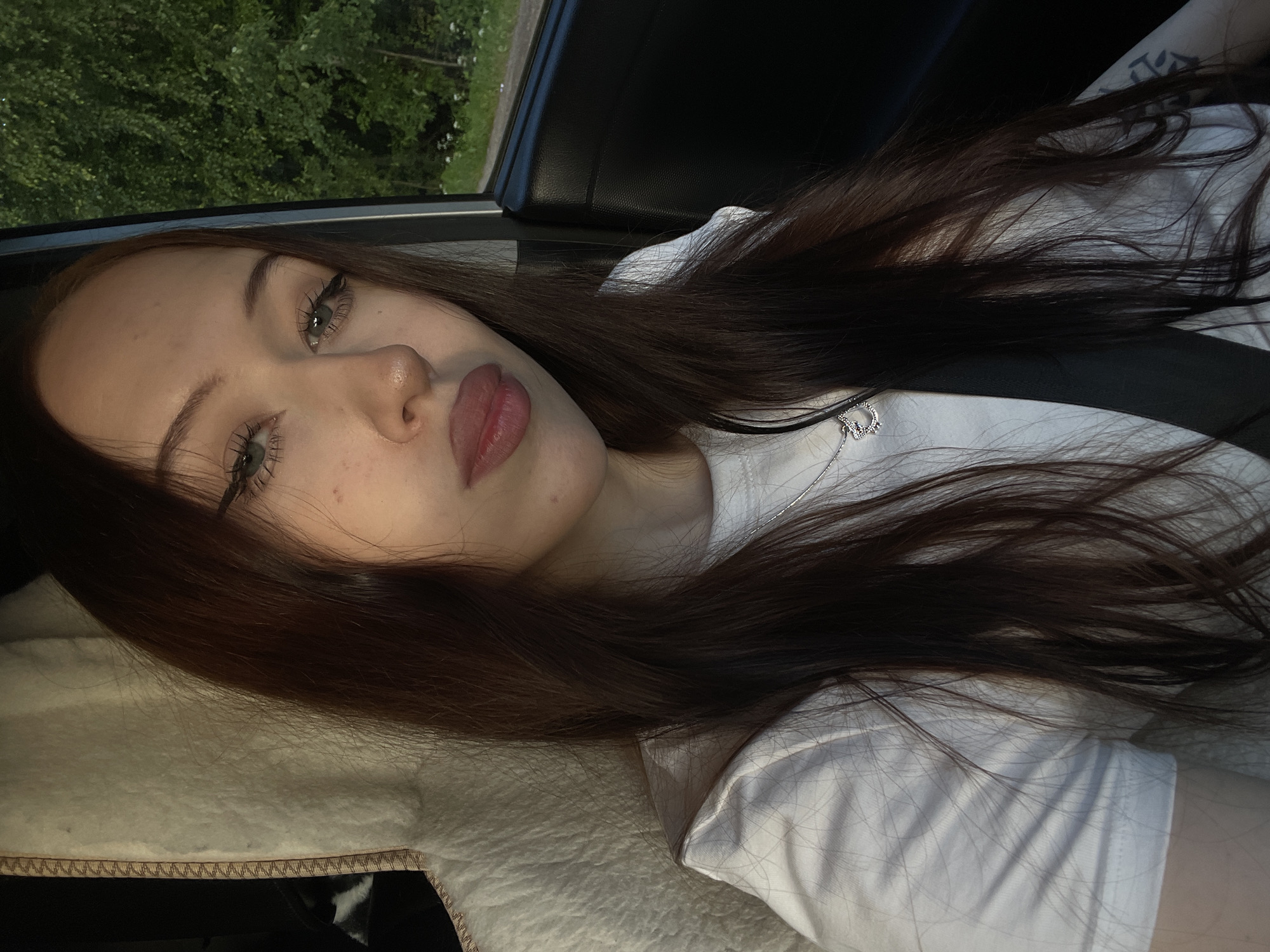 A young woman with long, dark brown hair is shown from the chest up, seated inside a vehicle. She is wearing a white t-shirt and a silver necklace. Her eyes are green, and she has noticeable eyeliner. The background shows trees and foliage outside the car window, suggesting an outdoor, possibly rural or suburban, setting. The interior of the car is visible, with a black car door on the right and a beige fuzzy seat cover on the left. A black seatbelt is visible across her chest. The image appears to be a selfie taken during the daytime. The location of Gavrilovka, Russia is not discernible from the visual information present in the image.