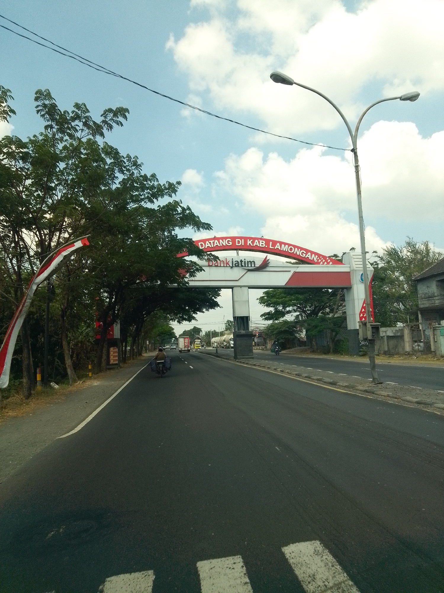 The image shows a view of a road in Sumurgenuk, Indonesia, as seen from a moving vehicle. A large archway spanning the road marks the entrance to "KAB. LAMONGAN" and features the logo of "bankjatim." The archway is red and white, with intricate patterns on the top red section. To the left of the road, there are trees and a white and red Indonesian flag banner. A streetlight with two lamps is positioned on the right side of the road. In the foreground, the asphalt road is visible with white pedestrian crossing markings. Further down the road, there are several vehicles, including cars, trucks, and a motorcycle carrying a person. The sky above is blue with scattered white clouds, and power lines are visible. The environment appears to be a typical roadside scene in Indonesia, possibly on a highway entering a regency.