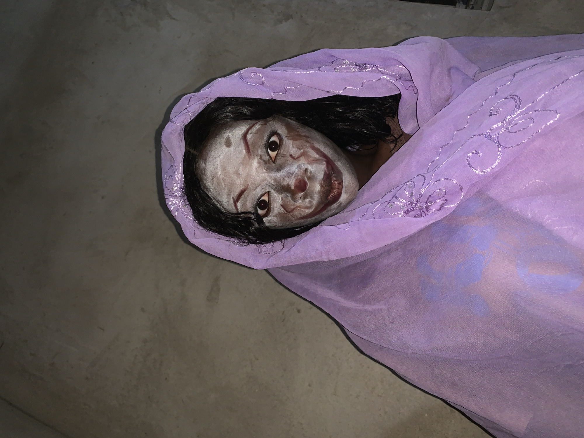 A person with theatrical makeup is lying on a gray concrete surface, wrapped in a light purple fabric with floral embroidery. The person's face is painted white, with dark markings around the eyes and mouth to exaggerate the features. Their hair is dark and wet. The setting appears to be indoors, with the concrete floor suggesting a utilitarian space. The exact location is Agban, Nigeria.
