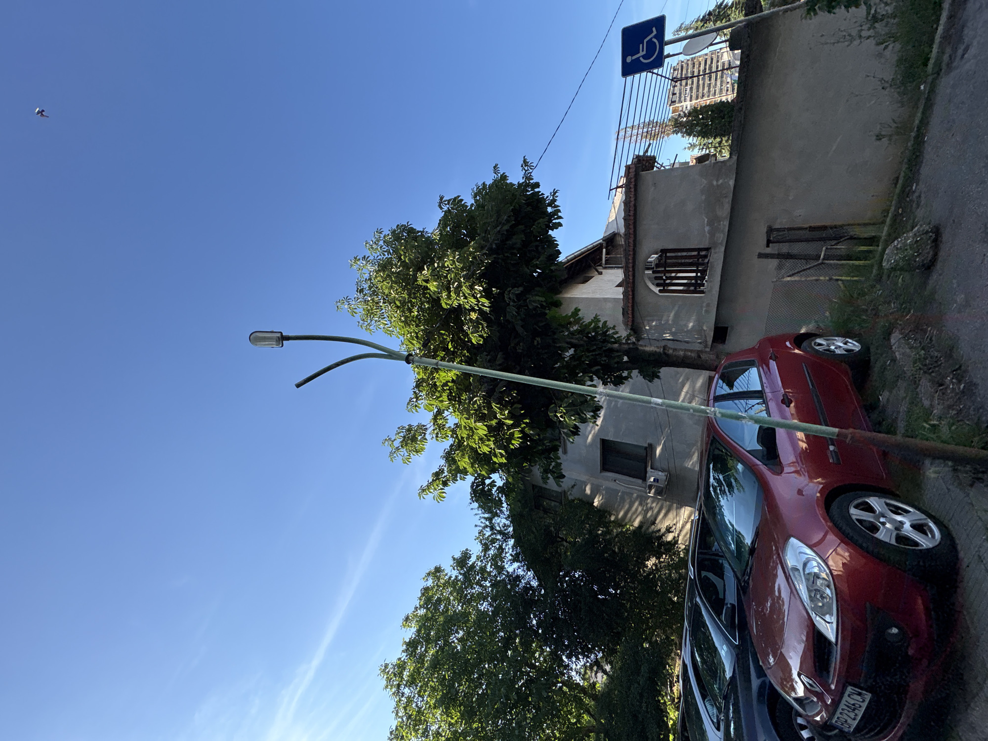 The image captures a street view in Vratsa, Bulgaria, under a clear blue sky with a few wispy clouds and a single bird flying. In the foreground, a red Renault Clio is parked, displaying the license plate "BP 2346 CM". To its left, a dark-colored car is partially visible. A street lamp, with a green pole and a modern lamp head, stands prominently in the center of the frame, angled towards the right. Behind the cars and lamp, a building with a grey facade, a tiled roof, and arched windows is visible. Trees with lush green foliage surround the building and extend to the left side of the image. A blue sign with a white wheelchair symbol, indicating accessibility, is mounted on a pole to the right of the building. The ground consists of pavement and a grassy area with rocks. In the background, beyond the building, a tall, modern apartment building and coniferous trees can be seen. The overall scene depicts an urban outdoor environment.