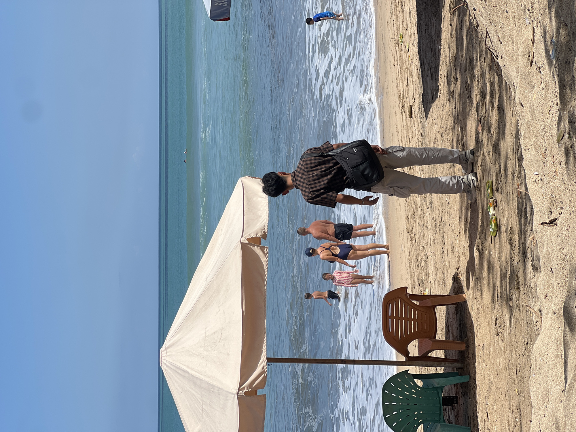 This is a photograph of a beach in Kuta, Indonesia. In the foreground, two empty plastic chairs, one green and one brown, sit on the sand. A man wearing a brown checkered shirt and light-colored cargo pants stands with his back to the camera, carrying a black shoulder bag. He is facing the ocean where several people are in the water. A large, light-colored umbrella provides shade in the left foreground. In the midground, a family with two children is wading in the shallow water. The father, wearing black shorts, stands to the right, while the mother, in a dark blue swimsuit and a blue cap, is to his left. A young boy is further out in the water to the left of the family, and a young girl in a pink and white patterned swimsuit stands between the parents and the boy. In the background, the turquoise ocean stretches to the horizon under a clear blue sky. Another umbrella, with red trim, is visible on the right side of the frame. The sand is light brown, with some debris scattered on it.