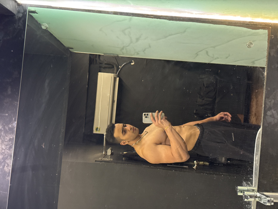 A person takes a selfie in a mirror, facing left. They are shirtless, wearing dark sweatpants, and sporting jewelry. The bathroom setting features a dark wall on the left, an AC unit, and light fixture above the mirror. The mirror has a cloudy, textured top. A bathroom sink is visible in the bottom right corner of the mirror's reflection.