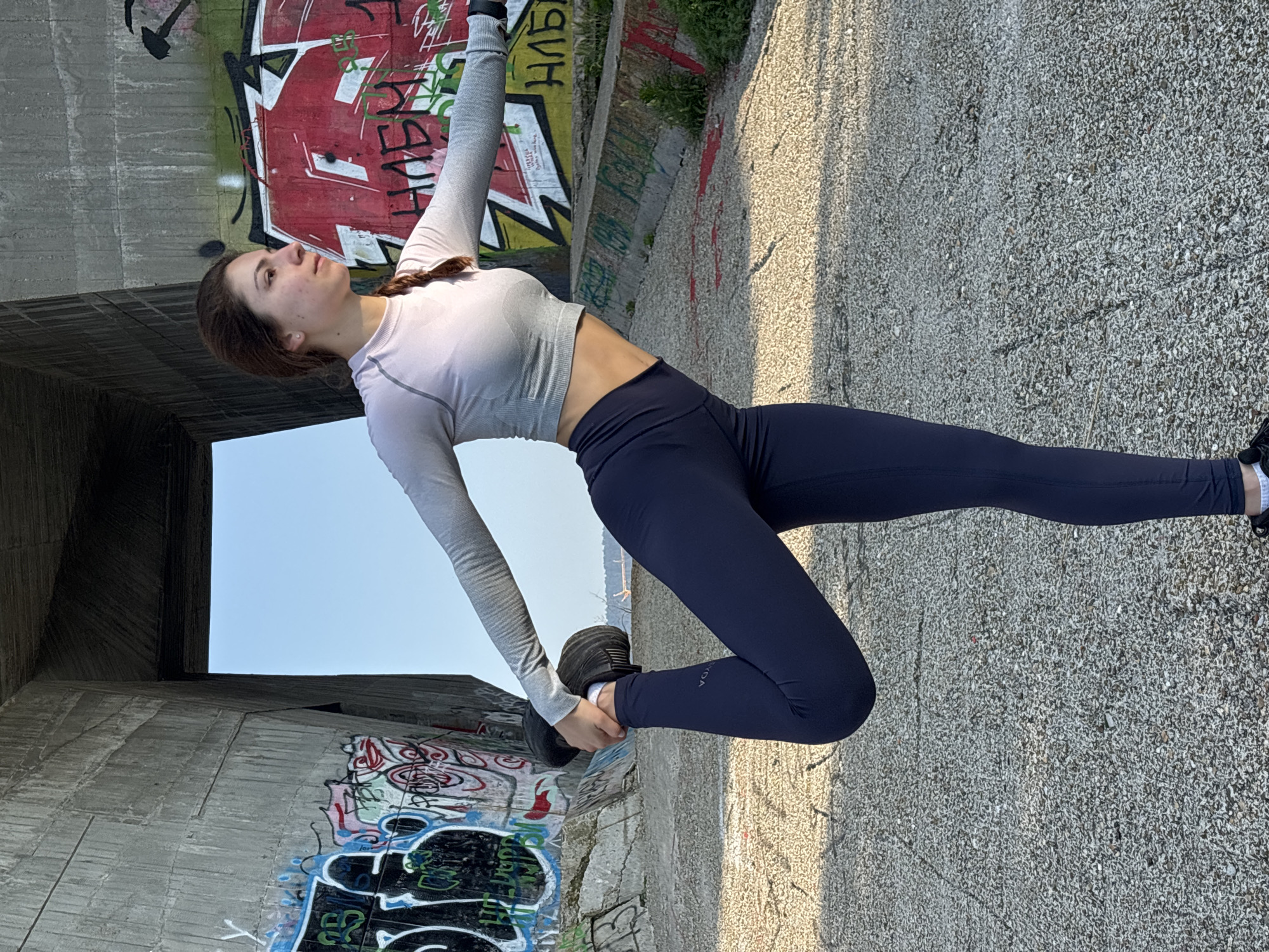 A young woman is captured in a graceful, extended stretching or yoga pose outdoors, likely beneath a concrete bridge or overpass in Varna, Bulgaria. She is balanced on her left leg, with her body arched backward, her left arm reaching upwards, and her right hand holding her right foot behind her, pulling it towards her head. Her long brown hair is styled in a braid, and her focused expression is directed upwards. She wears a form-fitting, light grey long-sleeved crop top and dark blue athletic leggings, with a small white text that appears to be "VIDA" visible on her right leg. Black athletic shoes and white socks complete her attire.
The setting is distinctly urban, featuring massive concrete walls and structures, characteristic of a bridge's underside, extensively covered in colorful graffiti. Among the spray-painted tags and designs, Cyrillic text can be seen, including "КГБ" (KGB) and "НАБ" (NAB) on the concrete surfaces. The ground is a light-colored, textured concrete or gravel. The scene is brightly lit by natural daylight, suggesting a clear, sunny day, with soft shadows indicating either morning or late afternoon. The overall atmosphere contrasts the woman's serene focus and the vibrant, rugged backdrop of the urban environment.