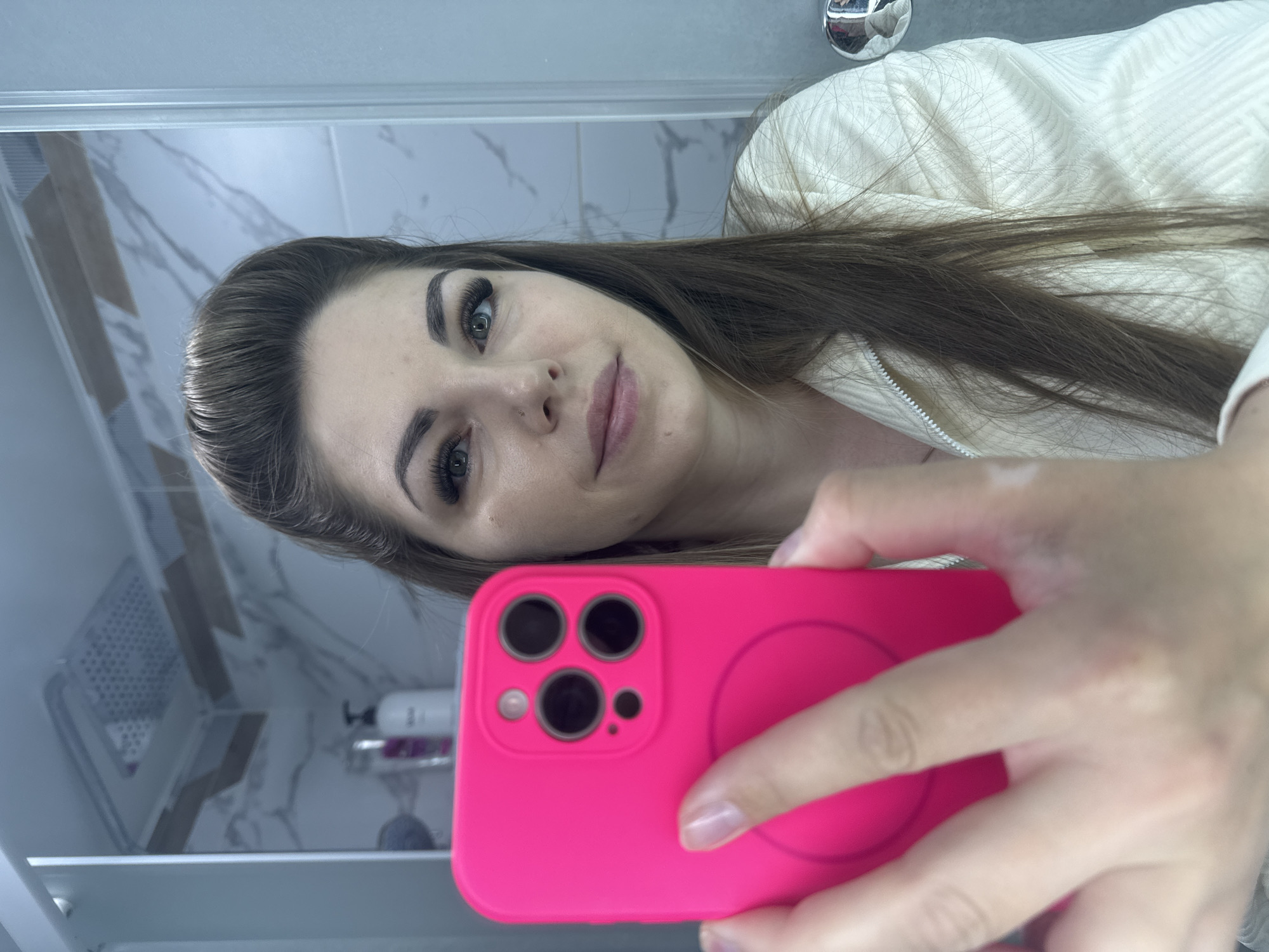 A person is taking a selfie in a bathroom. The individual has long brown hair, visible eyelashes, and light skin. They are wearing a white zip-up sweater. The phone is encased in a bright pink case. The bathroom has marble tile, a mirrored cabinet, and a stainless steel faucet. The location in the city of Раздолье, Russia is not discernible from the image.