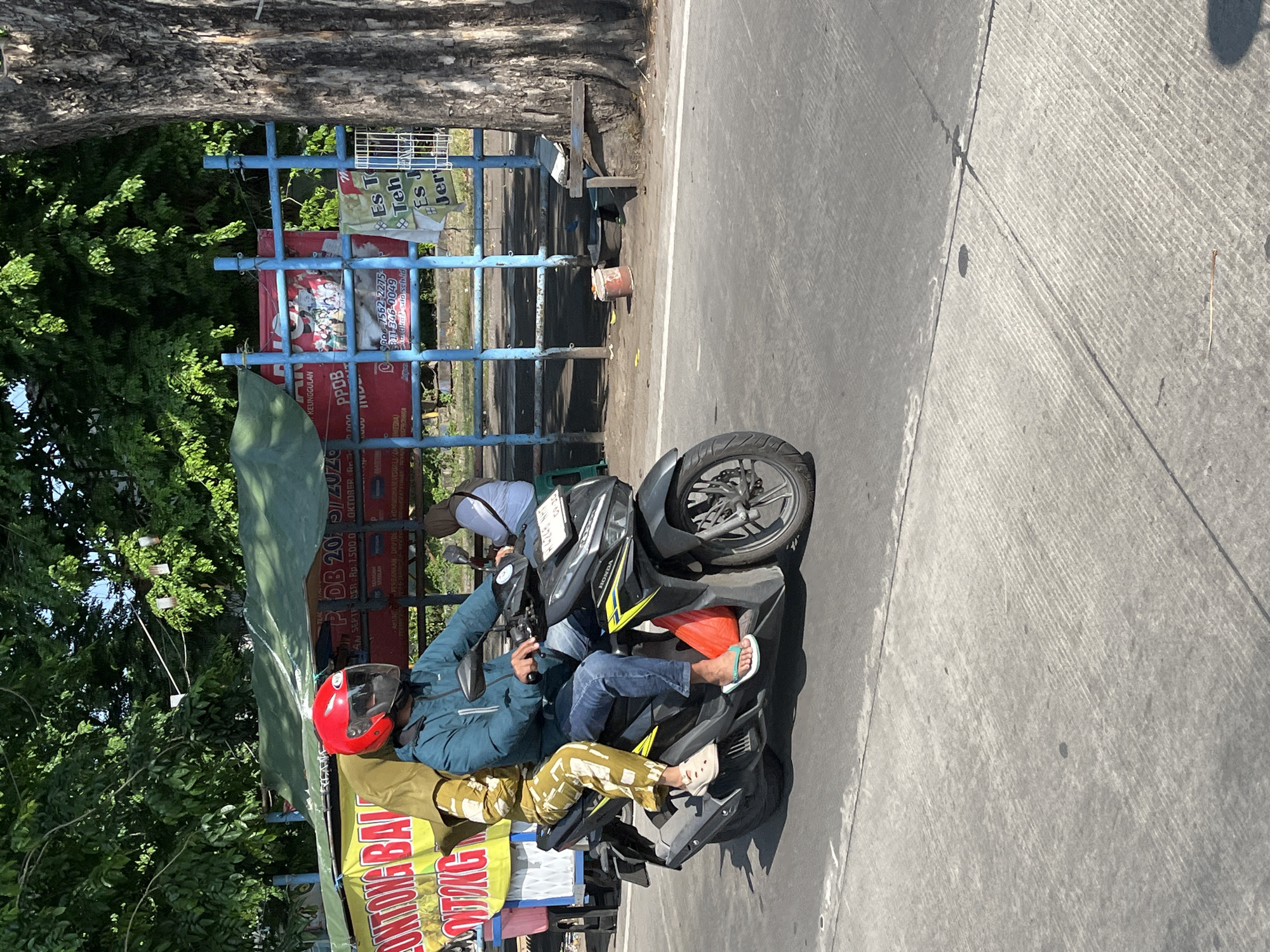 In Tulangan, Indonesia, a man wearing a teal jacket, blue jeans, and a red helmet is riding a black Honda motorcycle. A woman in a yellow and green patterned outfit and a yellow headscarf is a passenger on the motorcycle. They are riding on a paved road. Behind them, there is a large tree with rough bark and a blue metal fence with several signs. One sign is red and features a picture of people. Another sign is white and has text that reads "Es Teh Jer". There are also green tarps set up behind the fence, possibly for a market stall. Sunlight is casting shadows on the road and the surrounding area.
