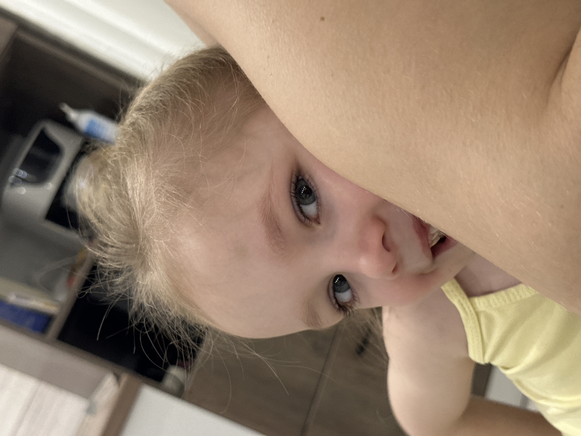 The image captures a candid, close-up moment featuring a young child, likely a toddler, peeking out from behind what appears to be an adult's arm or shoulder. The photo is taken from a slightly upward angle, with the child's head tilted, making the scene feel dynamic and playful.
The central subject is the child, who has light blonde hair, which appears fine and wispy, pulled back from their face. Their striking blue eyes are looking directly at the camera with an expression that suggests curiosity, playfulness, or perhaps mild shyness. The child's mouth is slightly open, revealing some teeth, indicating they might be about to smile, laugh, or speak. They are wearing a bright yellow top with a thin strap visible over their shoulder.
A prominent feature in the foreground and upper right of the image is the bare arm or shoulder of an adult, covering part of the child's face and creating a sense of intimacy or hiding. The adult's skin is light-toned with visible pores and fine hair, and a small, dark mole is present on the skin.
The background is largely out of focus, providing depth to the image but keeping the focus on the child. Blurry shapes suggest an indoor setting, possibly a domestic environment like a kitchen or living room, with dark furniture or cabinets and some lighter elements such as a counter or wall. A small, blurry white and blue object, possibly a bottle or container, is visible in the upper left background.
The lighting appears to be ambient indoor light, and there is no visible text in the image. The overall mood conveyed is one of warmth, affection, and a playful interaction between the child and the person holding them or taking the photo.