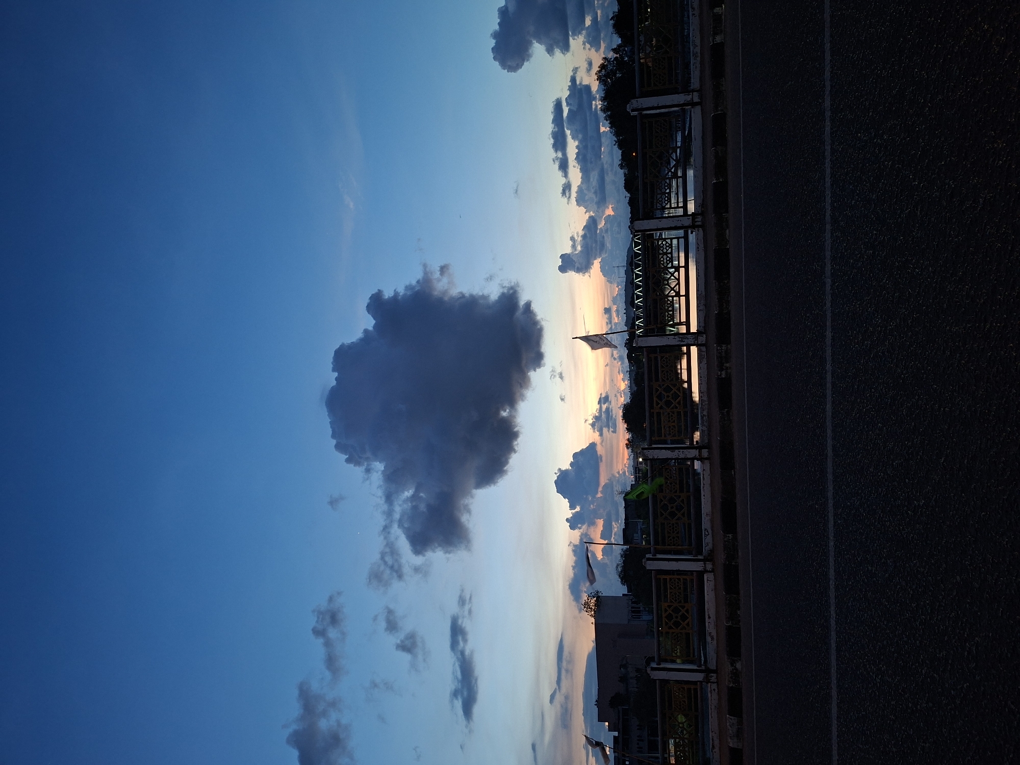 The image shows a view from a bridge or elevated structure, likely in Lhokseumawe, Indonesia, given the context. The sky transitions from a deep blue on the left to a lighter, sunset-tinged hue toward the center and right of the frame. There are scattered clouds throughout the sky, with a large, dark cumulonimbus cloud prominent on the left side. The bridge's structure, including railings and possibly walkways, are visible along the right side. A flag is visible flying over the bridge. A paved road is below. In the distance, one can discern buildings and trees silhouetted against the sunset sky.