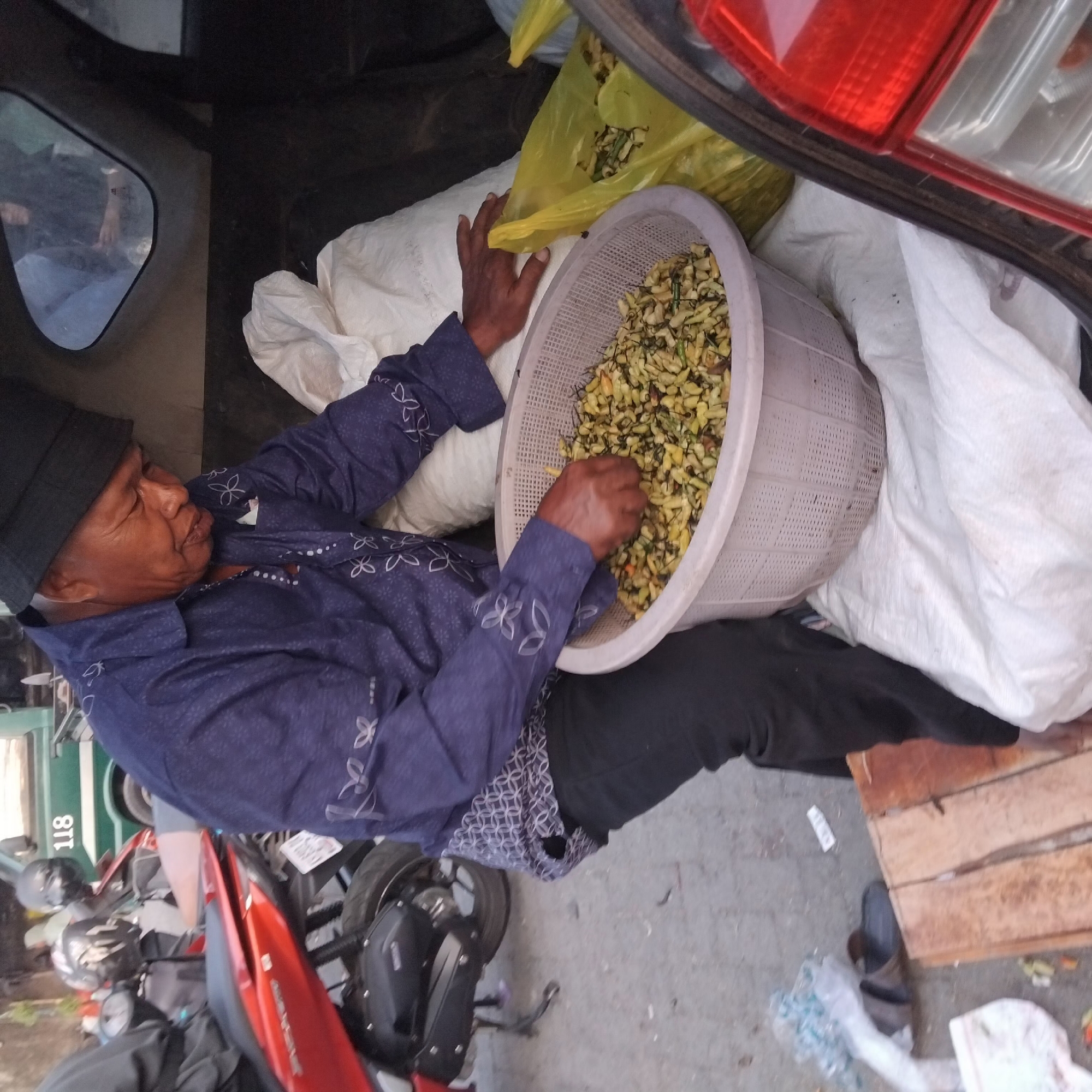 In Banjarbaru, Indonesia, an older man wearing a black bucket hat and a dark blue patterned shirt with white floral designs is sitting in the open back of a vehicle, sorting through a large, light-colored plastic basket filled with what appears to be food items, possibly chili peppers or similar produce. His hands are actively engaged in separating and arranging the contents of the basket. To his right, a bright yellow plastic bag is partially visible, also containing produce. The vehicle appears to be parked outdoors, on a street with a paved surface, and motorcycles are visible in the background, suggesting a typical urban or semi-urban Indonesian setting. Other items, such as white sacks and a wooden crate, are also present in the vehicle's cargo area. The overall scene depicts a moment of preparation or packaging of goods, likely for sale or transport.