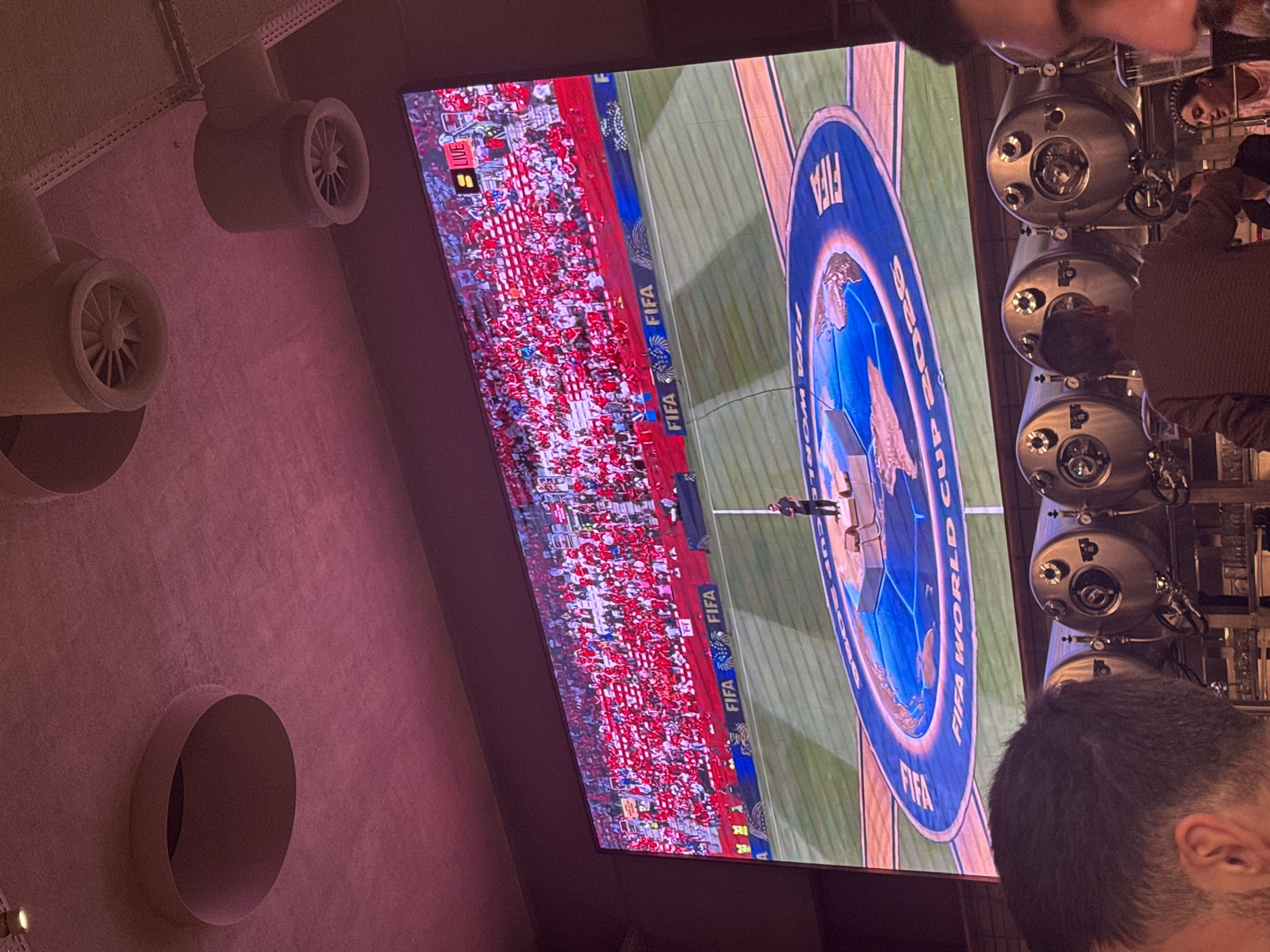 The image shows a large screen displaying a FIFA World Cup broadcast, with a stadium full of fans in red attire. A single person stands on a stage in the center of the screen, in front of a circular graphic with a world map. The screen is illuminated by pink lighting. In the foreground, several people are visible, including a person whose head and ear are in the lower left corner, and another person in the lower right corner, only their nose and mouth are visible. Behind them, several large, silver beer tanks are lined up. The scene appears to be in a bar or restaurant, with exposed ceiling elements and lighting fixtures visible at the top of the image.