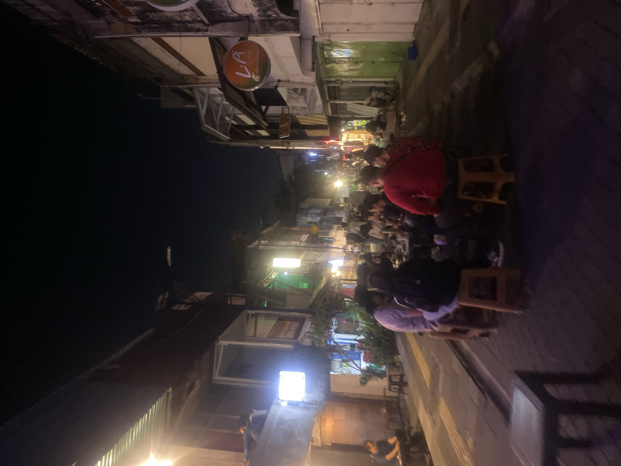 This is a nighttime outdoor scene in Banjarmasin, Indonesia, likely a street or alleyway lined with buildings that appear to be shops or restaurants. Several groups of people are seated at tables, engaged in conversation and dining. The lighting is dim, with some artificial lights illuminating the area. One prominent sign features the text "LA ICE MANGOSOURT". The overall atmosphere suggests a casual, lively evening gathering.