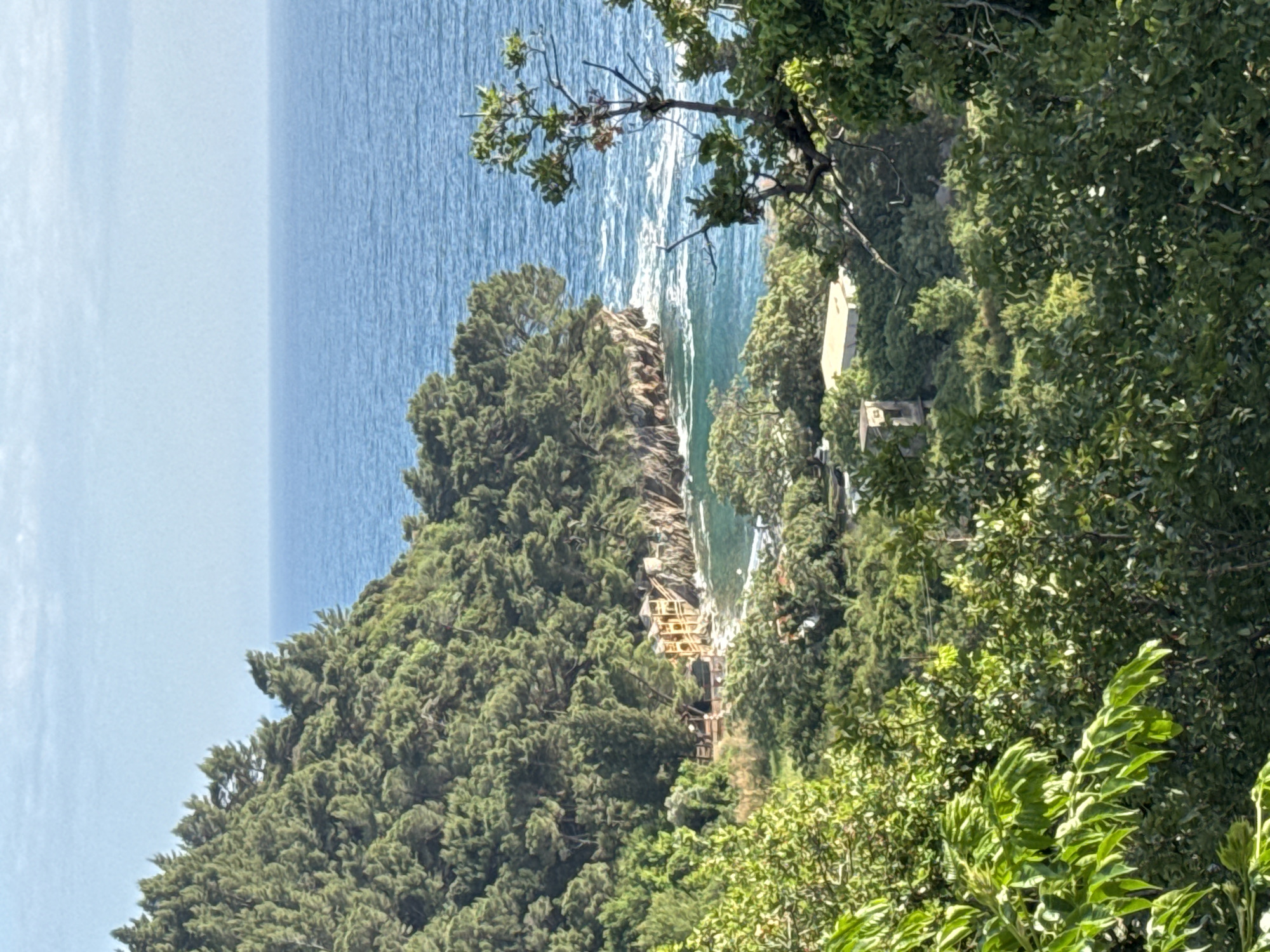 The image captures a coastal scene in Brežine, Montenegro. The sea occupies the upper left of the frame. The coastline is characterized by a cliff face and foliage. A structure, scaffolding, can be seen. Lush green trees fill the right side of the image.