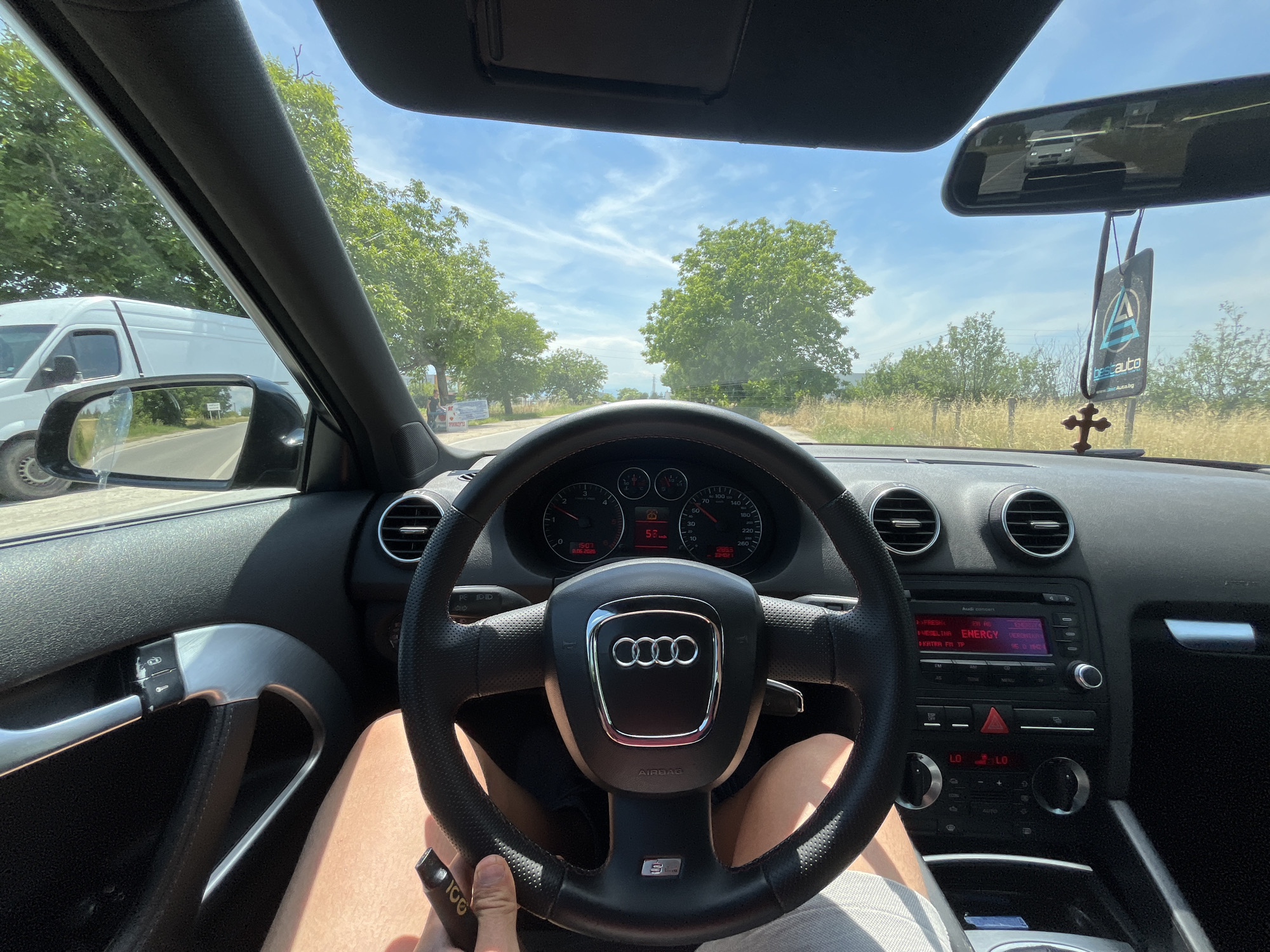 The image is a point-of-view shot from inside a car, likely an Audi, driving on a road outside the city of Skutare, Bulgaria. The driver's legs are visible, and they are holding a car key. The steering wheel, dashboard with gauges, and part of the car's interior are in focus. Outside the car, through the windshield and side windows, a rural landscape with trees and a bright blue sky with clouds is visible. A white van is seen in the side mirror. A air freshener and a wooden cross are hanging from the rearview mirror. The road appears to be paved and leads through a grassy and tree-lined area.