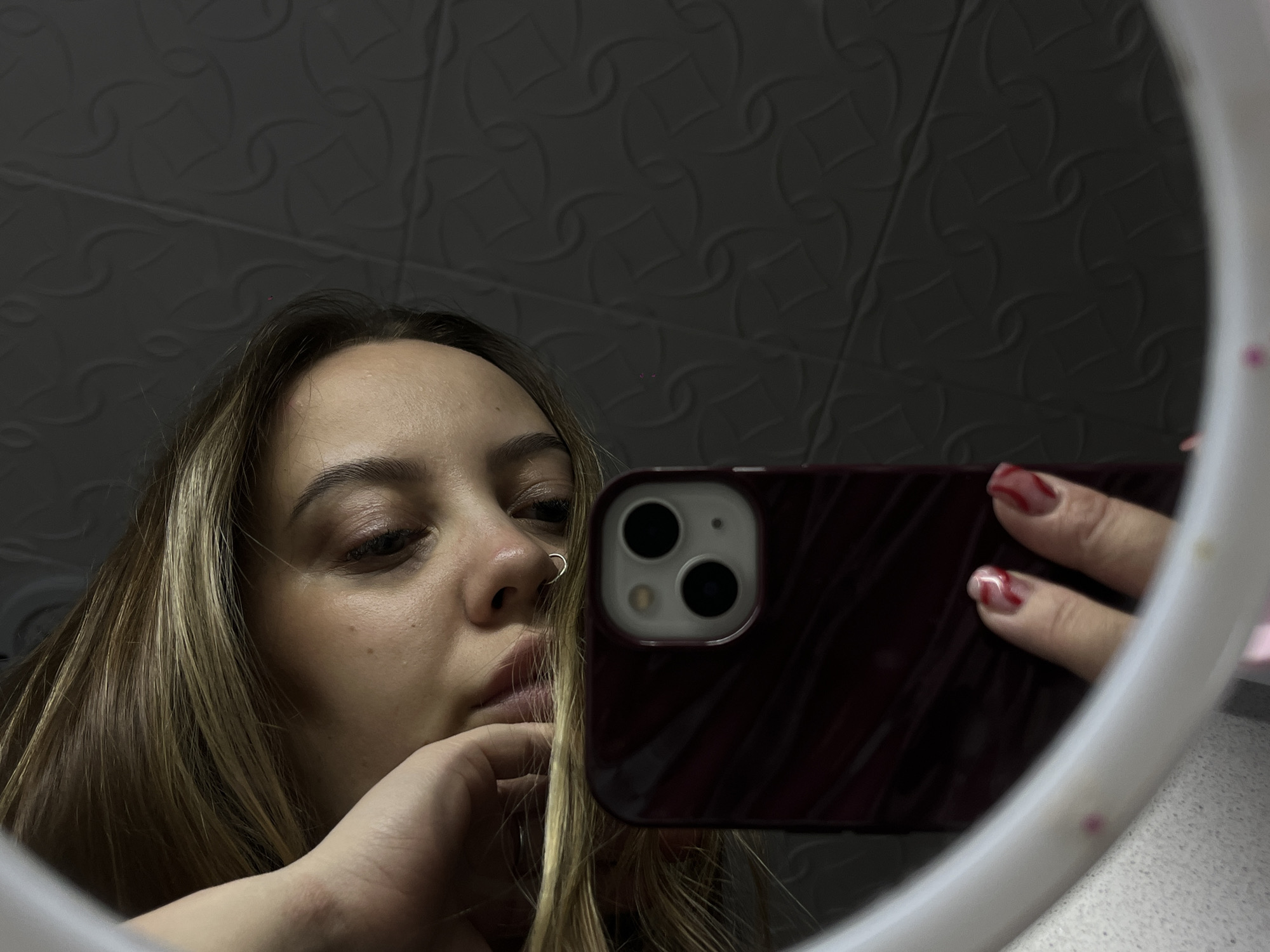 A woman is taking a selfie in a mirror. She has blonde hair and is wearing a nose ring. Her right hand is raised to her chin. Her left hand is visible holding a phone with a dark red case. The background is a textured grey wall. There are no indications that this photo was taken in Novopokrovskaya, Russia.