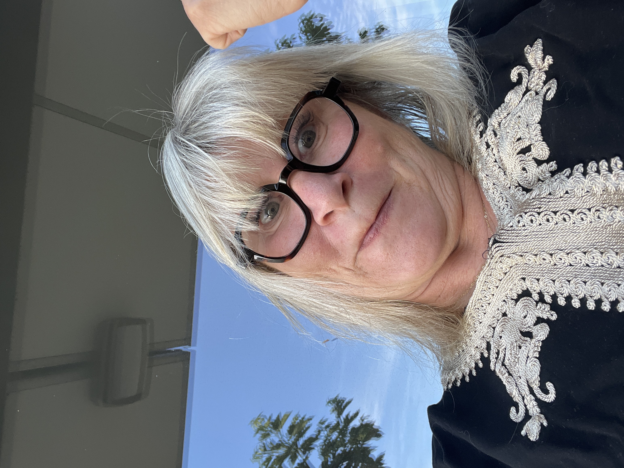 A close-up, tilted portrait captures an individual, likely female, with light grey and blonde hair and prominent black-framed glasses. She appears to be looking slightly off-camera with a neutral, contemplative expression. Her attire consists of a dark garment, possibly black, adorned with intricate silver or white embroidered patterns around the neckline and upper chest area, suggesting a formal or culturally significant piece of clothing.
The background is divided, with a clear, bright blue sky visible on the right side, featuring reflections of palm trees, indicating a sunny outdoor setting or a reflection off a large window. On the left, a darker, possibly indoor or shaded, grey surface is visible, showing a subtle reflection or outline of a rectangular object. A person's hand is partially visible in the upper right corner, possibly holding the camera for a selfie. The image conveys a daytime scene with clear weather conditions. This scene is situated in Chambœuf, France.