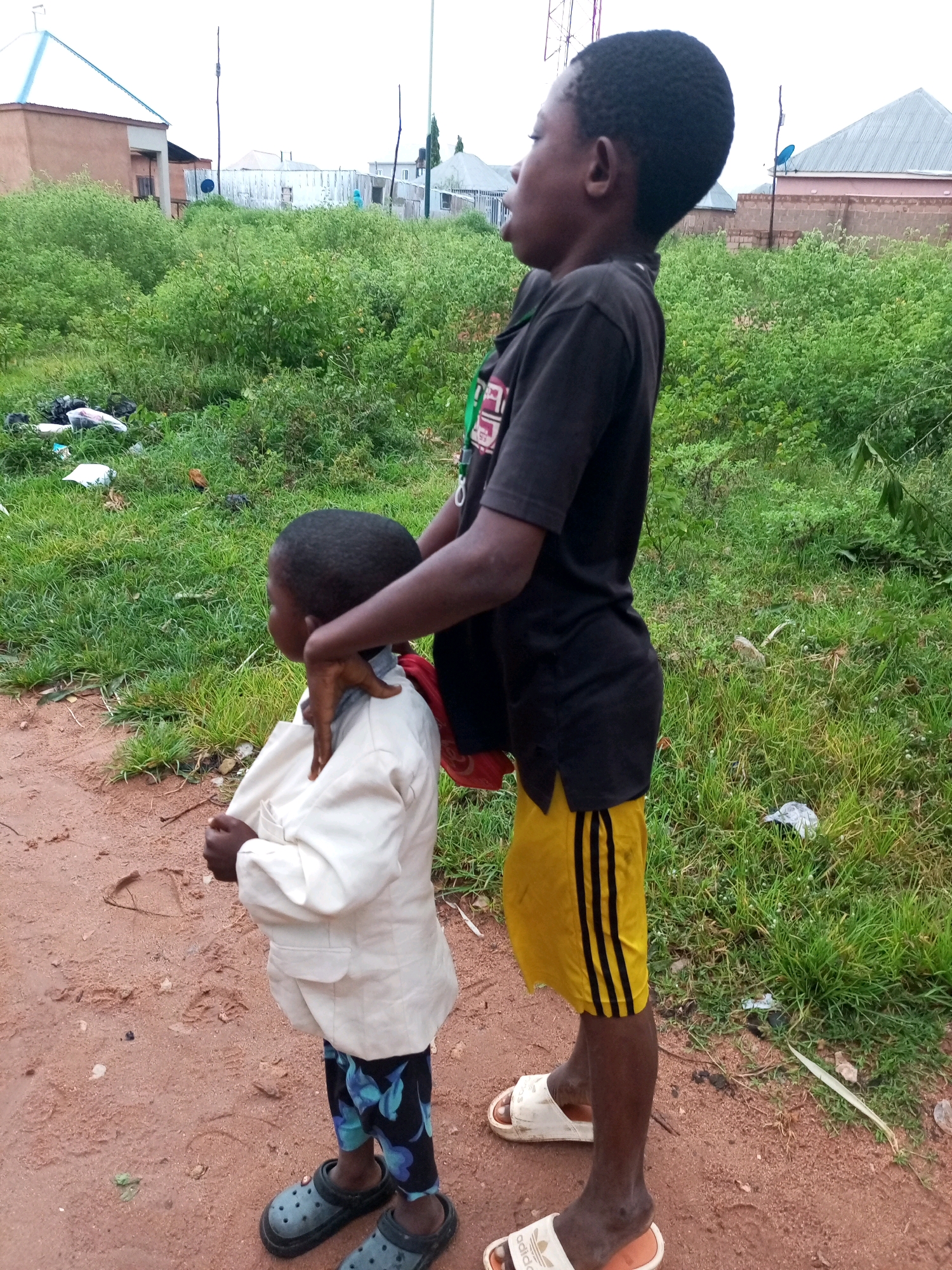 The image shows two young boys standing in an outdoor environment. The boy in the foreground is wearing a light-colored jacket, dark patterned pants, and dark clogs. He is looking to his left. The taller boy behind him is wearing a black t-shirt and yellow shorts with black stripes. He is looking to his right and has his hand on the neck of the boy in front of him. The background consists of overgrown green vegetation, some scattered debris, and buildings in the distance, suggesting a semi-urban or rural setting within Gombe, Nigeria. The ground beneath the boys is dirt and appears somewhat muddy, with visible footprints. The sky is overcast.