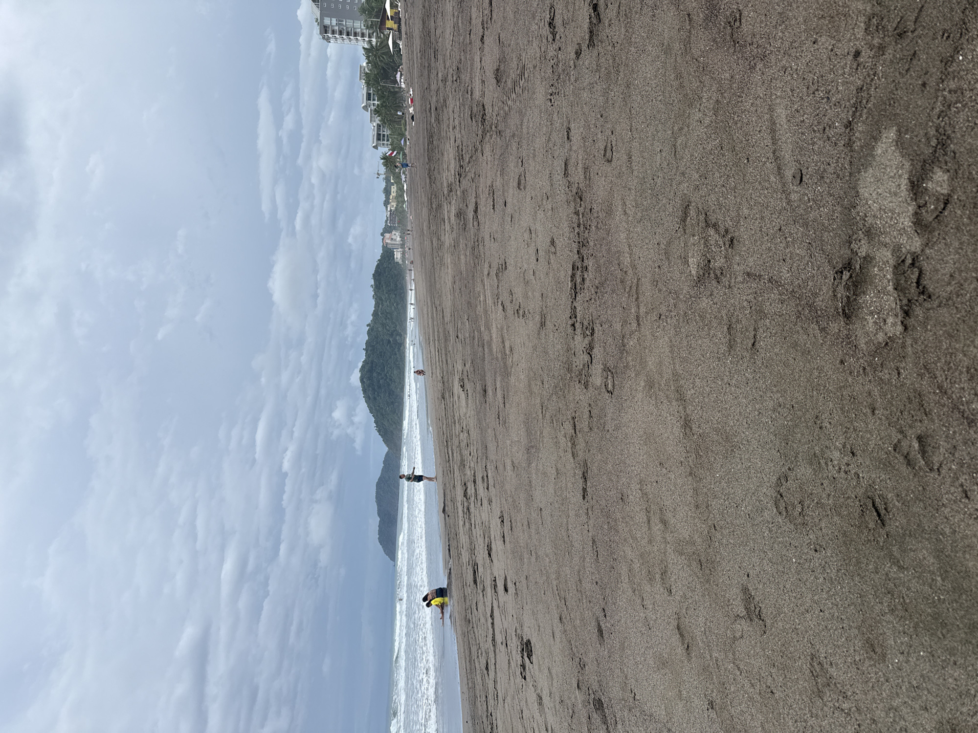 The image depicts a wide, sandy beach in Jacó, Costa Rica, under a cloudy sky. The dark sand is marked with numerous footprints. In the foreground, the sand is granular and slightly damp in places.
Further out, near the water's edge, several people are present. Two individuals, one in a yellow top and another in a darker garment, are seated in the shallow water, appearing to rest or converse. A lone figure, wearing a green shirt and dark shorts, stands on the sand near the water, looking towards the ocean. A small child is visible in the distance on the right, near the water.
The ocean exhibits moderate waves, with whitecaps breaking near the shore. In the background, a line of lush green hills rises, forming a natural backdrop to the beach. Beyond the hills, several multi-story buildings, likely hotels or condominiums, are visible along the coastline, indicating the presence of a developed resort area. White umbrellas and some beach chairs are scattered on the sand in front of these buildings. The overall atmosphere suggests a typical, overcast day at a popular beach destination.