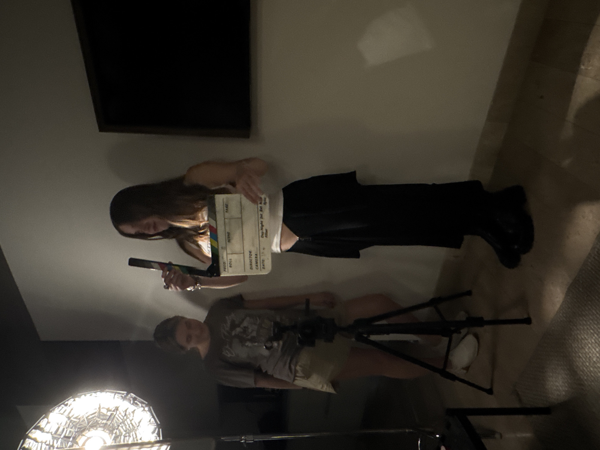 The image shows an indoor film set with two people. One person, seen from the torso up, is holding a clapperboard open. This person has long, brown hair, wears a white top, and black bottoms. The second person stands next to a tripod-mounted camera. This person wears a t-shirt and shorts and white shoes. The scene is lit by a bright lamp, and there is a black rectangle, likely a piece of set design, hanging on the wall. It is not possible to determine the specific location within Mexico City from the image.
