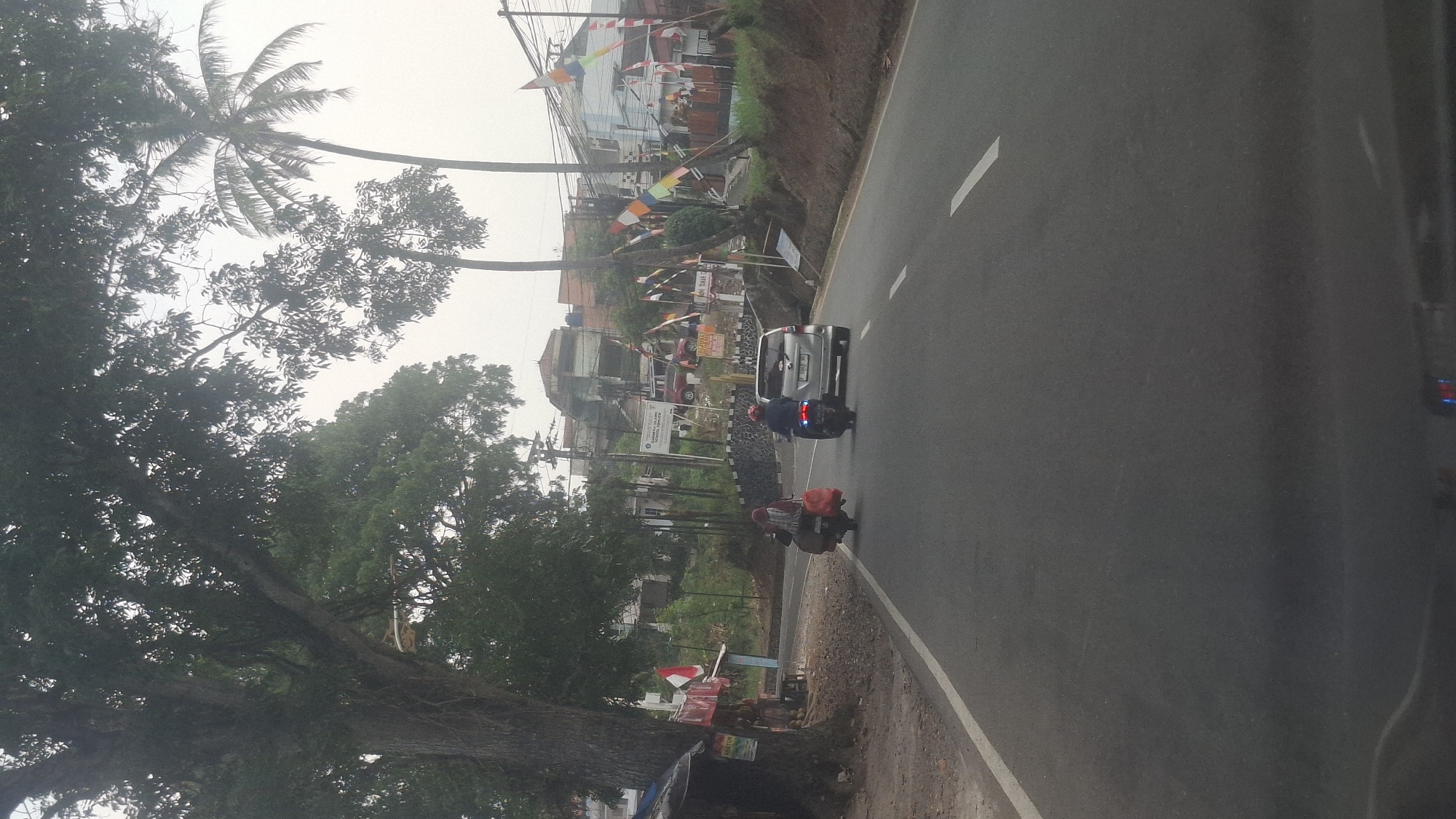 The image depicts a scene on a paved road in Cilaku, Indonesia, with a backdrop of lush vegetation and buildings. In the foreground, a silver car is visible, followed by a motorcycle carrying a rider wearing a red helmet. Further ahead, another motorcycle is seen with a rider draped in a patterned cloth, transporting bags. The road is lined with trees, including a large, mature tree on the left side and palm trees on the right. Buildings and houses are scattered on a hillside in the background, some adorned with Indonesian flags. The sky is overcast, suggesting either early morning or late afternoon. The overall impression is a snapshot of daily life on a road in a tropical urban or semi-urban setting.