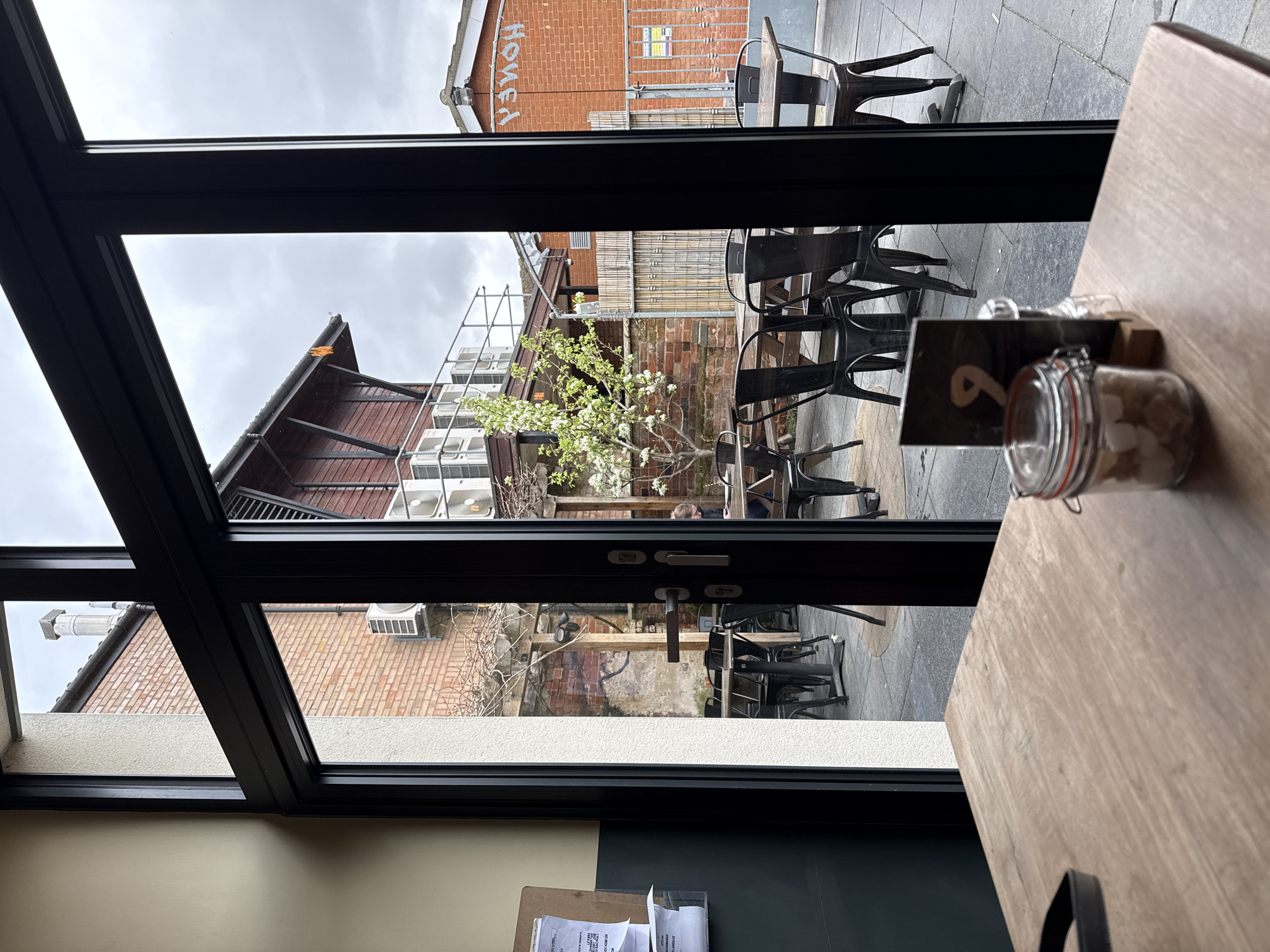The image is taken from an indoor vantage point, looking out through a black-framed window. The scene presents an outdoor patio area. Outside, there are multiple metal chairs and tables arranged, suggesting a seating area for a café or restaurant. In the background, buildings constructed of red brick are visible. The word "Honey" is painted on the side of the brick building, and scaffolding is set up against it. A blossoming tree adds a touch of nature to the urban scene. Inside, a wooden table is partially visible, with a jar containing small, white objects, and a table number holder. The setting suggests a location in an urban area in East Suffolk, United Kingdom, most likely a café or restaurant with an outdoor seating area.