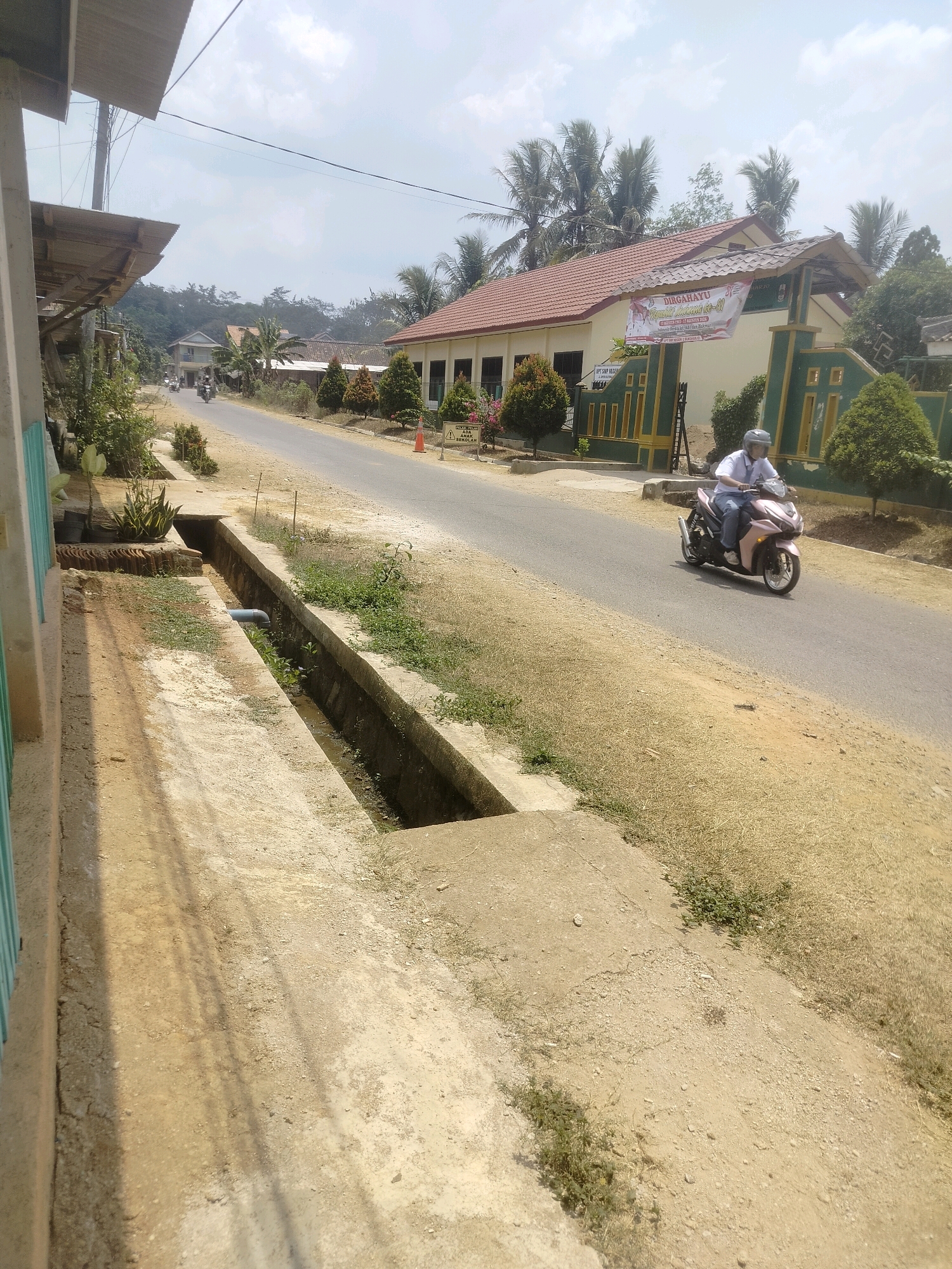 In the city of Rejosari, Indonesia, a motorcycle rider, wearing a white shirt and helmet, drives down a paved road. The rider is heading away from the viewer, towards a building with a red-tiled roof and a banner that reads "DIRGAHAYU REPUBLIK INDONESIA KE-77" in Indonesian. The building appears to be a school, as indicated by a sign that says "Waspada Anak Sekolah" (Beware School Children). The road is bordered by dry, dusty earth and a concrete drainage ditch on the left side of the image. Scattered trees and vegetation are visible, along with a few other distant structures. The sky is partly cloudy.