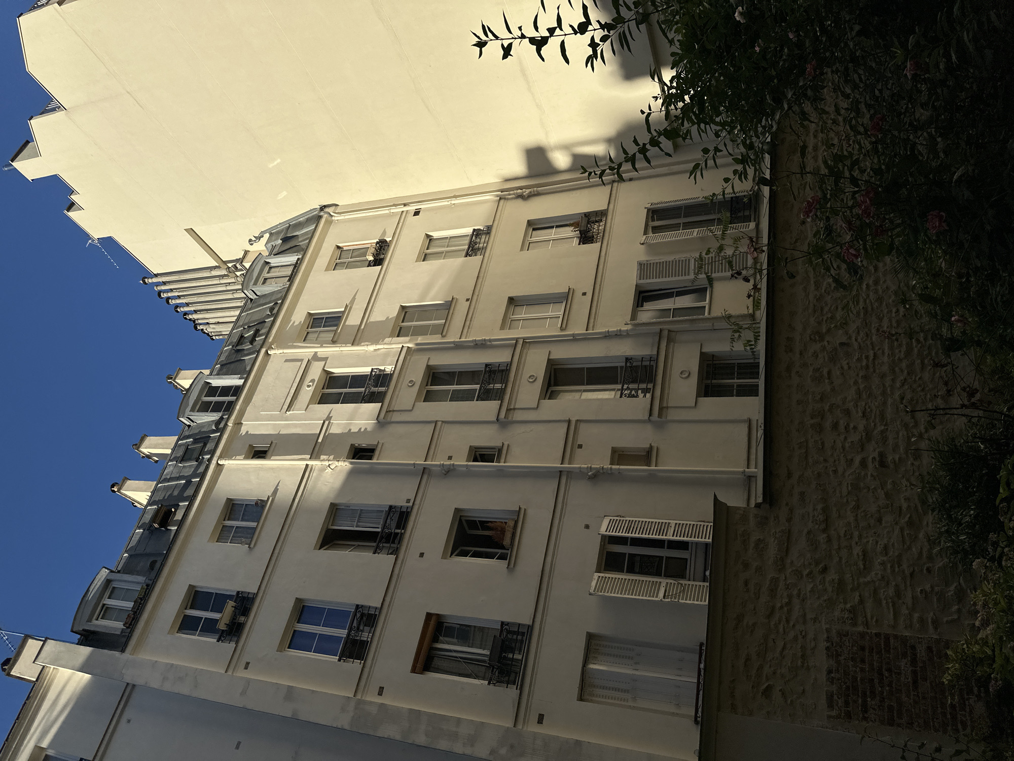 A low-angle shot captures a cream-colored multi-story building with a mansard roof against a clear blue sky in Paris, France. The building features numerous windows, some with open shutters, and decorative iron railings. Shadows from an adjacent building are cast on the facade of the building on the right. In the foreground, a stone wall borders a dark garden area with some foliage and pink flowers. The scene appears to be a residential building, possibly an apartment building, given the architectural style.