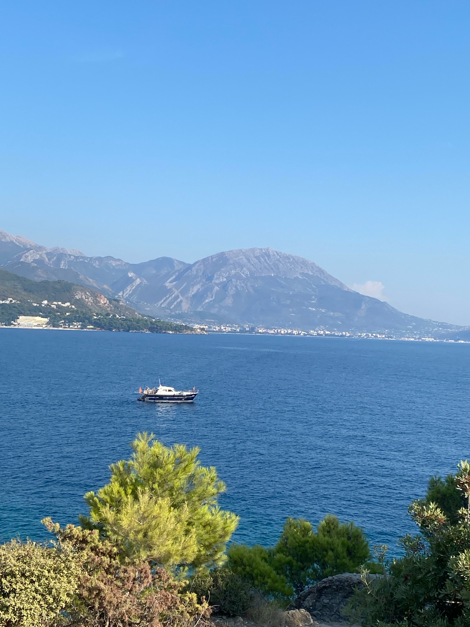 The image shows a serene view of the Adriatic Sea near Sutomore, Montenegro, under a clear blue sky. In the middle ground, a small, dark-hulled motorboat with a white superstructure is sailing on the calm, blue water. The boat appears to be carrying a few individuals. In the background, a coastal town with white buildings is visible nestled at the base of rugged, grey mountains. Lush green vegetation, including trees and bushes, occupies the foreground, framing the scenic vista.
