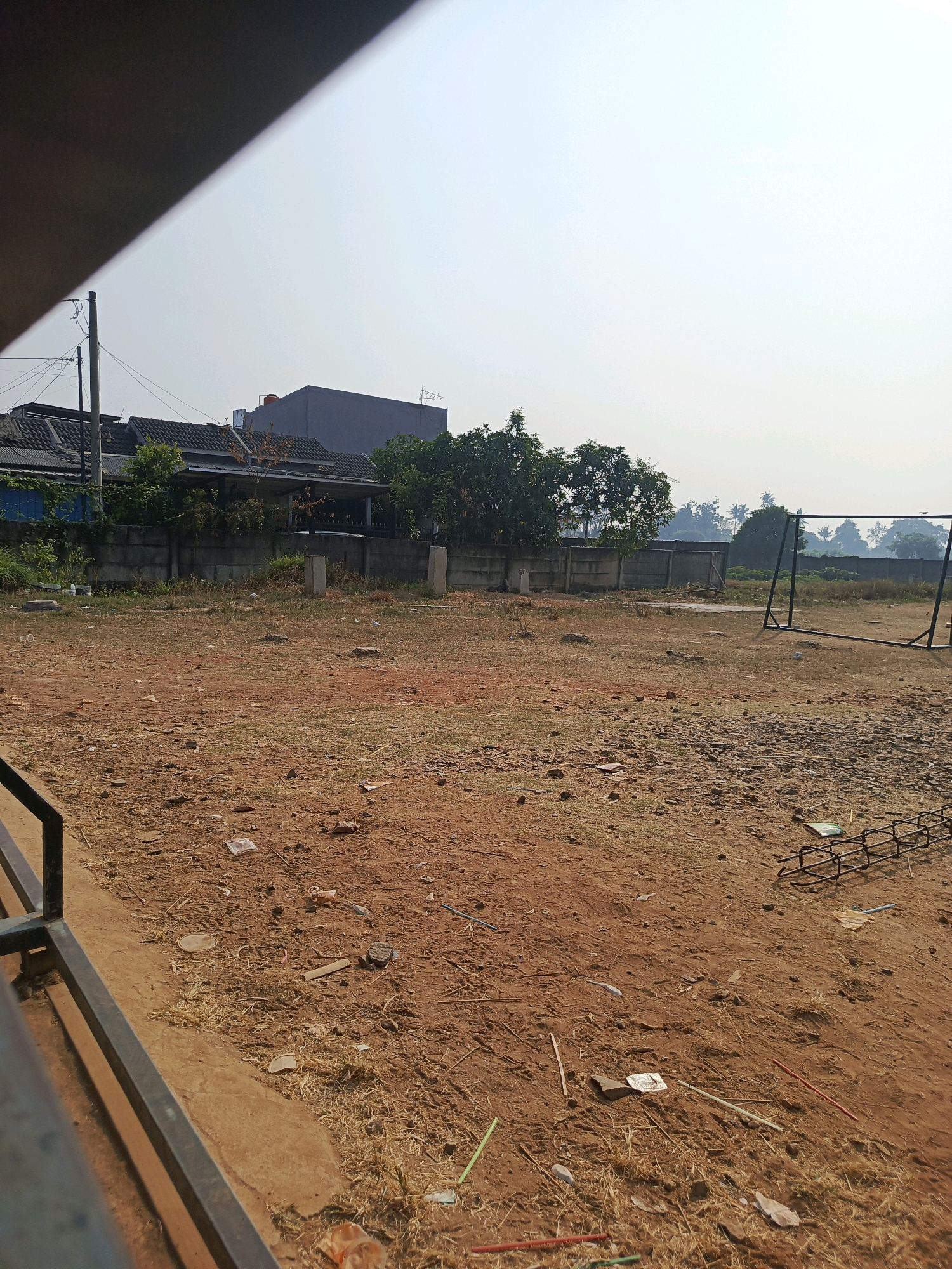 The image depicts an outdoor scene in Jati Mulya, Indonesia, characterized by a dirt field with sparse dry grass and scattered debris. In the background, a concrete wall separates the field from a residential area with several houses, some featuring tiled roofs and antennas. Trees are visible behind the wall, and in the far distance, more trees and a hazy sky suggest a humid or polluted atmosphere. On the right side of the field, a black metal structure, possibly part of a goal frame for a sport, is present. In the foreground, a dark metal railing suggests the edge of a platform or structure from which the photo was taken. The ground is predominantly brown earth with small rocks and dried vegetation.