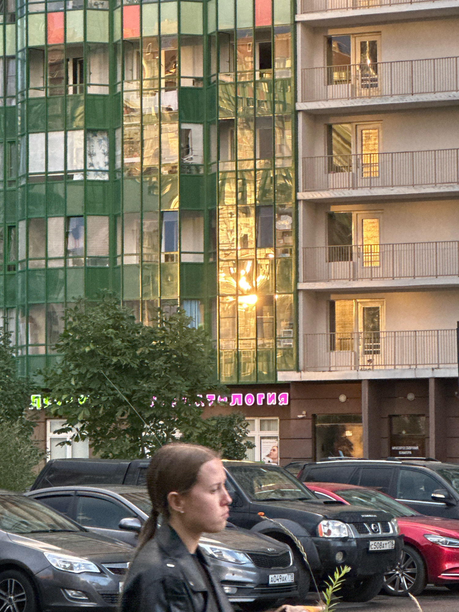 The image shows a street scene in a city, possibly Russia, with a modern building featuring a green glass facade and balconies. The sun is setting or rising, casting a warm glow on the building's windows, creating reflections of trees and the sky. In the foreground, several cars are parked, and a young woman with her hair in a ponytail is standing and looking to the right. The overall atmosphere is calm and urban.
