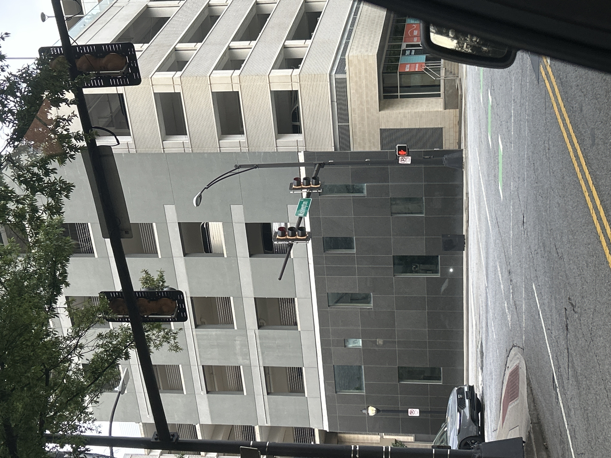In an urban environment, a gray multi-story parking garage with white horizontal accents is visible. A portion of a gray building with dark gray tiled lower floors is also present. Traffic lights and street signs are mounted on poles. One street sign reads "Washington." A pedestrian signal displays a red walking figure, indicating "Don't Walk." A silver car is parked on the left side of the frame. The ground is an asphalt road with white lane markings and some green painted markings. Trees with green leaves overhang the scene. A reflection of the surroundings is visible in the side mirror of a vehicle from which the photo is taken.