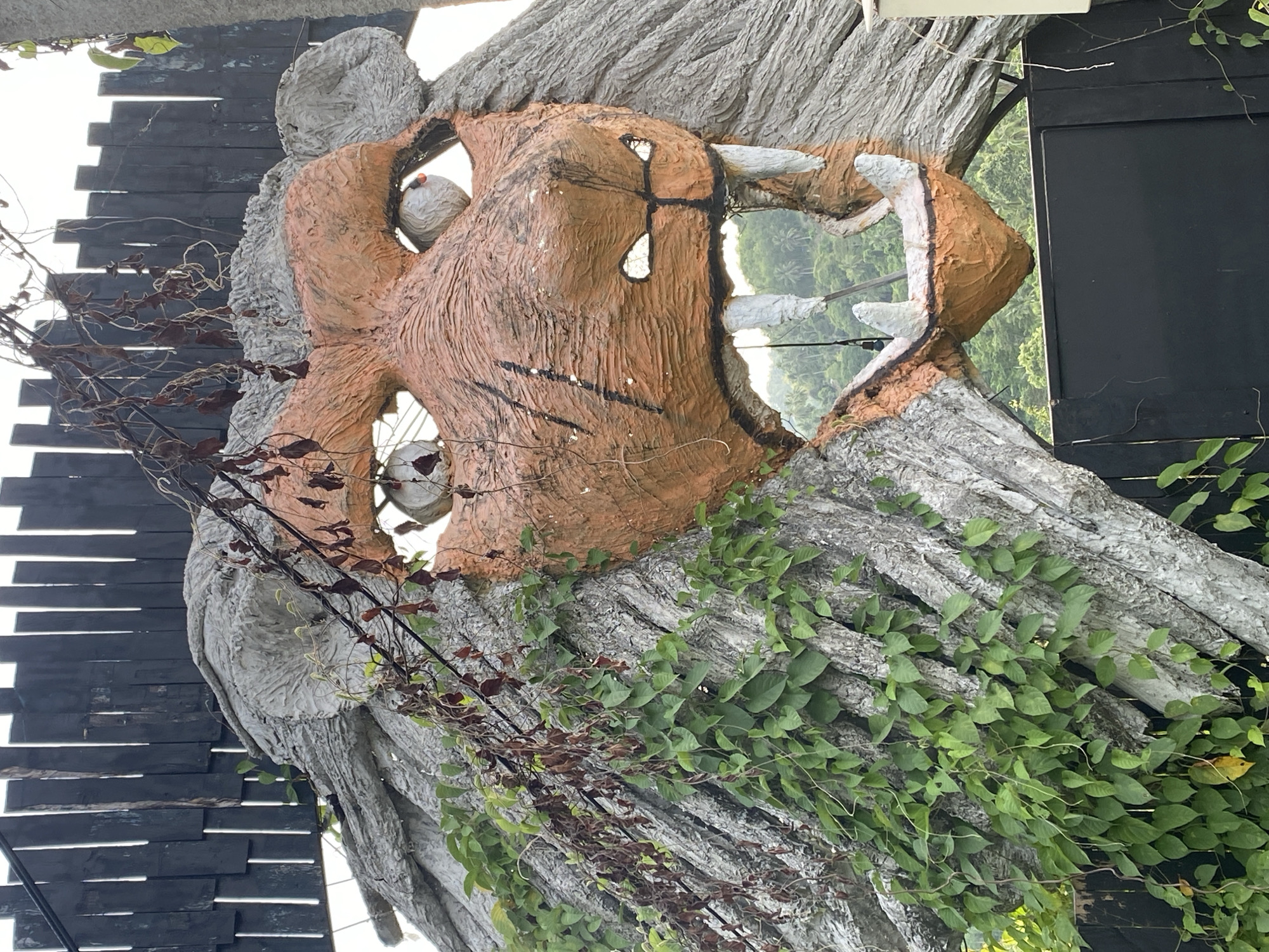A close-up of an outdoor art installation in Moo 2, Thailand, shows a sculpted tiger head emerging from a simulated tree trunk. The tiger's head is rendered in orange and brown hues with exaggerated features, including large teeth and black markings. The surrounding artificial tree trunk is textured in gray, with green vines climbing over it. A black wall with staggered wooden slats is visible in the background, along with a glimpse of green foliage through an opening in the tiger's head.