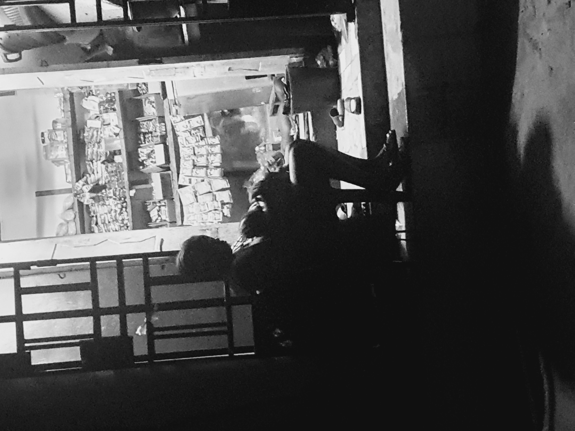 The image shows a person sitting on the street in front of a store in Lokoja, Nigeria. The store appears to be stocked with various goods. The person is dark and hard to see and could be on a porch. A grid railing is near the person.