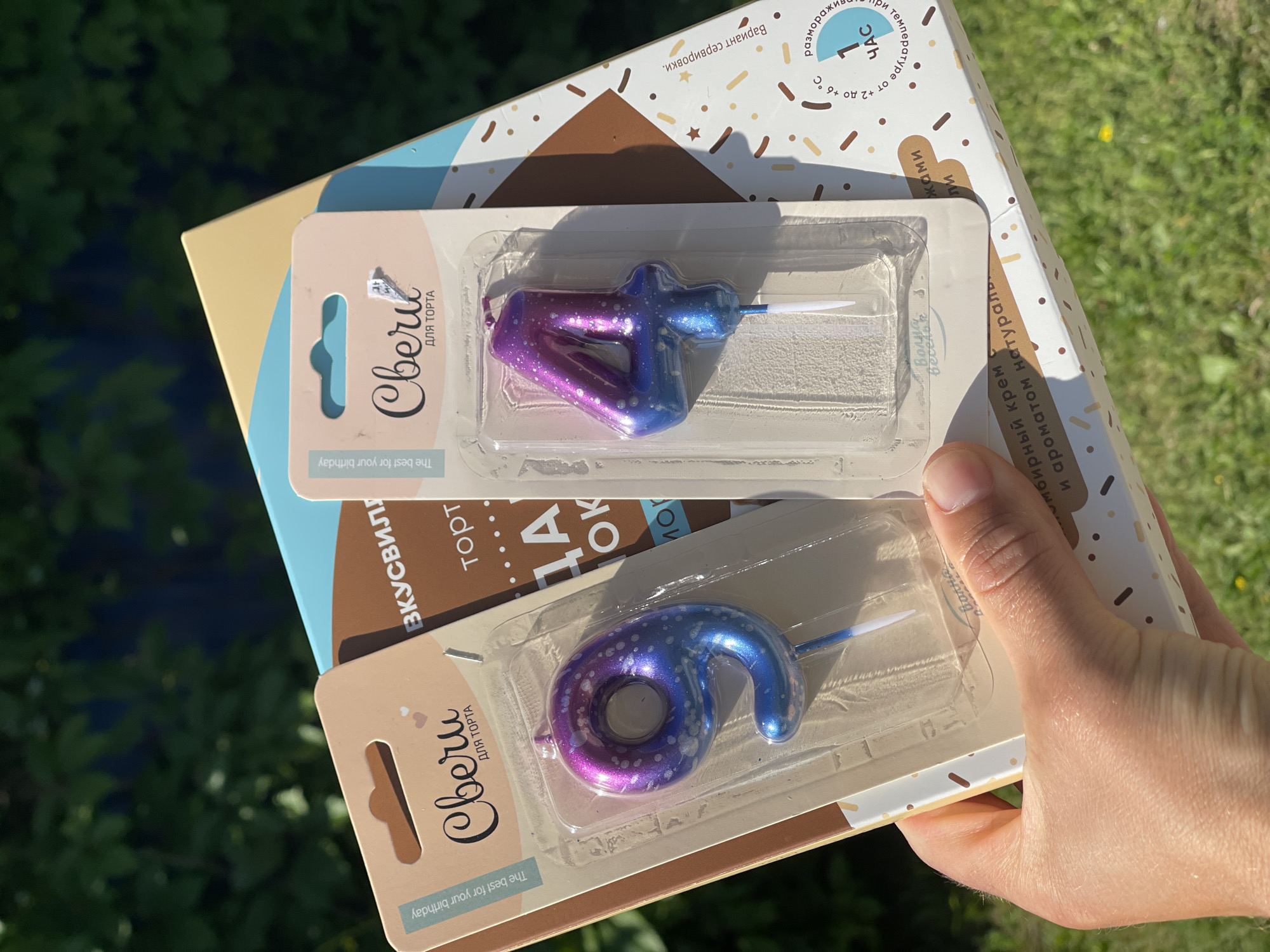 In an outdoor setting, a person's hand holds two birthday cake candles shaped like the numbers nine and four, packaged in clear plastic. The candles have a gradient color of purple and blue with white speckles. The "9" candle is on the left, and the "4" candle is on the right. Behind the candles is a box with a patterned design and some text. The background is blurred green foliage, suggesting trees or bushes.