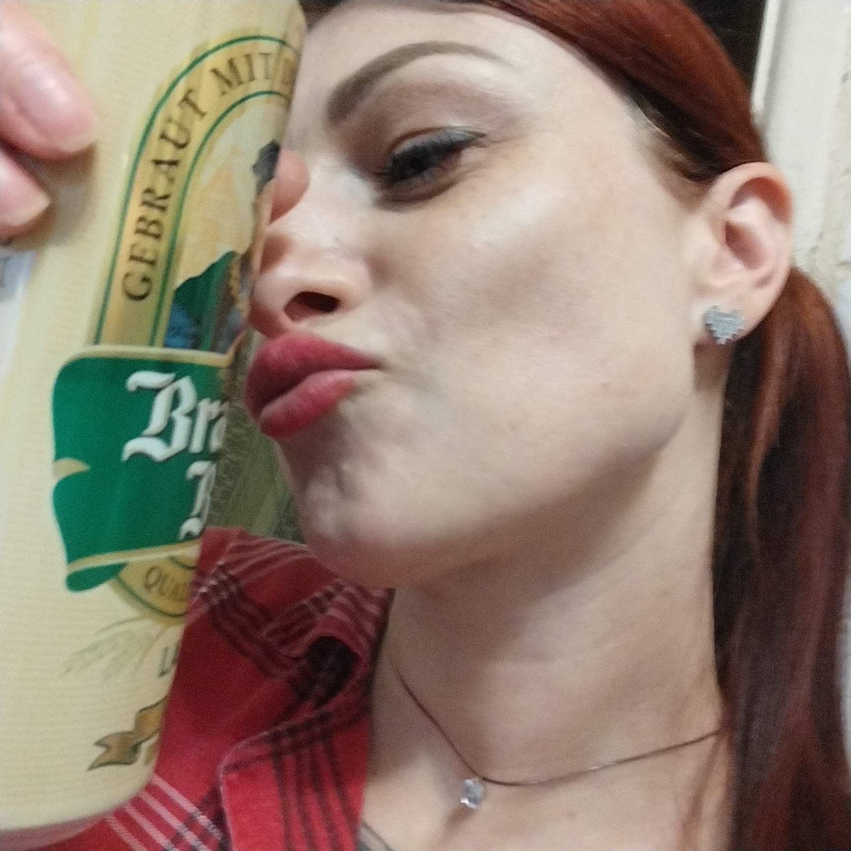 A woman with red hair and a red plaid shirt is holding a beer can to her face. She is wearing a small heart-shaped earring and a thin necklace with a pendant. The beer can has text that appears to be in German, with words like "GEBRAUT" and "MIT". The background is out of focus, but appears to be a light-colored wall. The image was taken in Забалоцкі сельскі Савет, Belarus.