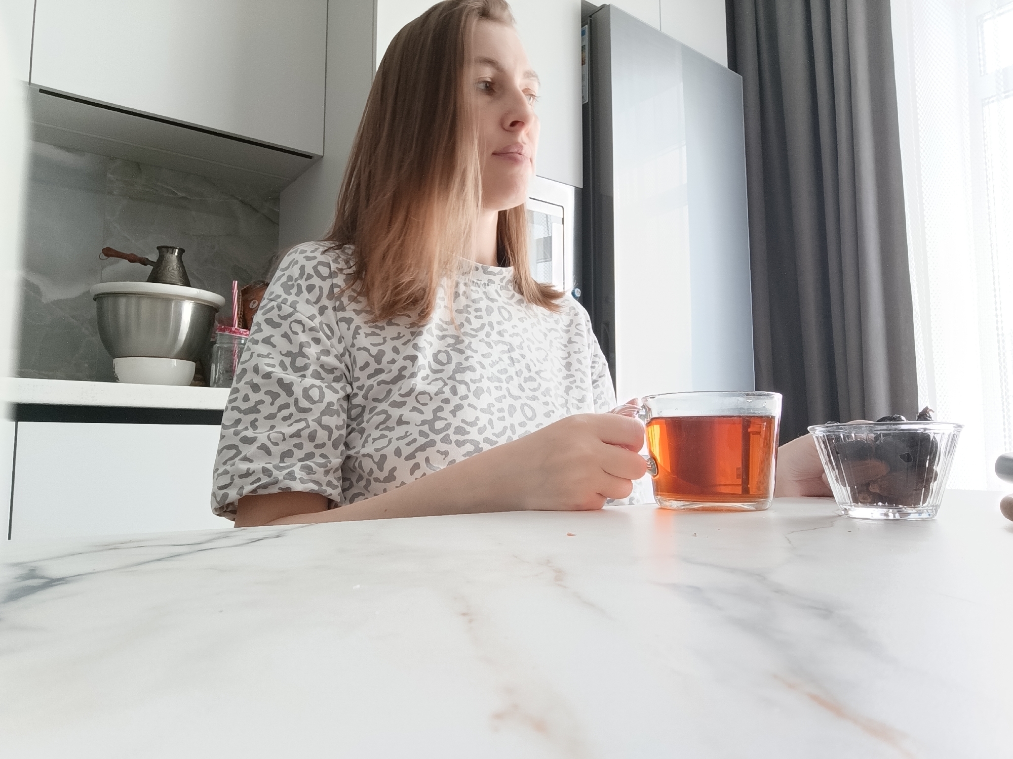 This image depicts an indoor scene within a modern residential kitchen or dining area, located in Chaikovsky, Russia. A female individual, characterized by shoulder-length light brown hair and a light-colored top with a grey leopard-print pattern, is seated at a white table featuring grey marble-like veining. Her right hand grasps a clear glass mug filled with a reddish-brown liquid, identified as tea. A small, clear, ribbed glass bowl containing dark dried fruits or nuts is positioned on the table to her right. The subject's gaze is directed off-camera to her right. The background on the left reveals white overhead kitchen cabinets, a grey marble-patterned backsplash, a silver metal mixing bowl, and a metal cezve (Turkish coffee pot) with a wooden handle. On the right side of the background, a silver refrigerator and a dark grey curtain partially obscuring a brightly lit window, behind which a white sheer curtain is visible, complete the scene.