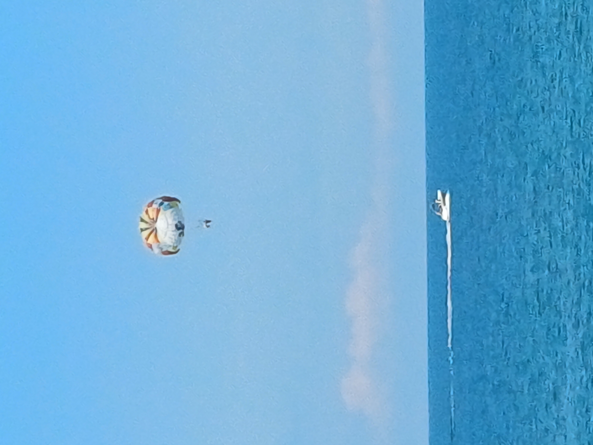 An aerial view captures a vibrant parasail, adorned with colorful stripes, suspended against a clear blue sky. Attached to the parasail by thin ropes, a person is being towed through the air. Below, a white speedboat races across the turquoise waters of the sea, leaving a foamy white wake in its path. The horizon line, where the sea meets the sky, is faintly marked by a thin strip of white cloud. The scene is set in the city of Sochi, Russia, a renowned resort town on the Black Sea coast.