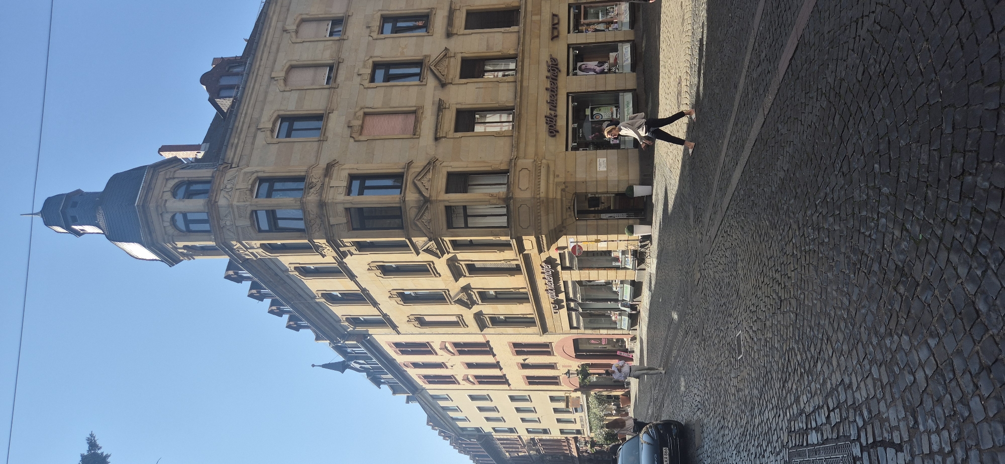 The image depicts a sun-drenched street scene in Mainz, Germany, specifically the picturesque cobblestone street in front of the Optik Niederhofer store. The architectural style of the buildings suggests a historic European city, likely from the late 19th or early 20th century, characterized by ornate facades, bay windows, and mansard roofs.
In the foreground, a black car is parked on the left side of the street. Two women are walking on the cobblestone street. One woman, wearing a light-colored top and dark pants, is walking towards the right of the frame. Another woman, wearing a light-colored top and dark pants, is walking in the same direction. The woman on the right is further into the frame and appears to be crossing the street.
The buildings lining the street house various businesses, with signage visible for "Optik Niederhofer" which specializes in optical goods. The storefronts have large display windows showcasing products. The street itself is paved with cobblestones, a common feature in many European cities, and shows signs of wear and tear. The sky is clear and blue, indicating fair weather. The overall impression is one of a vibrant urban environment with a blend of historical architecture and modern commercial activity.