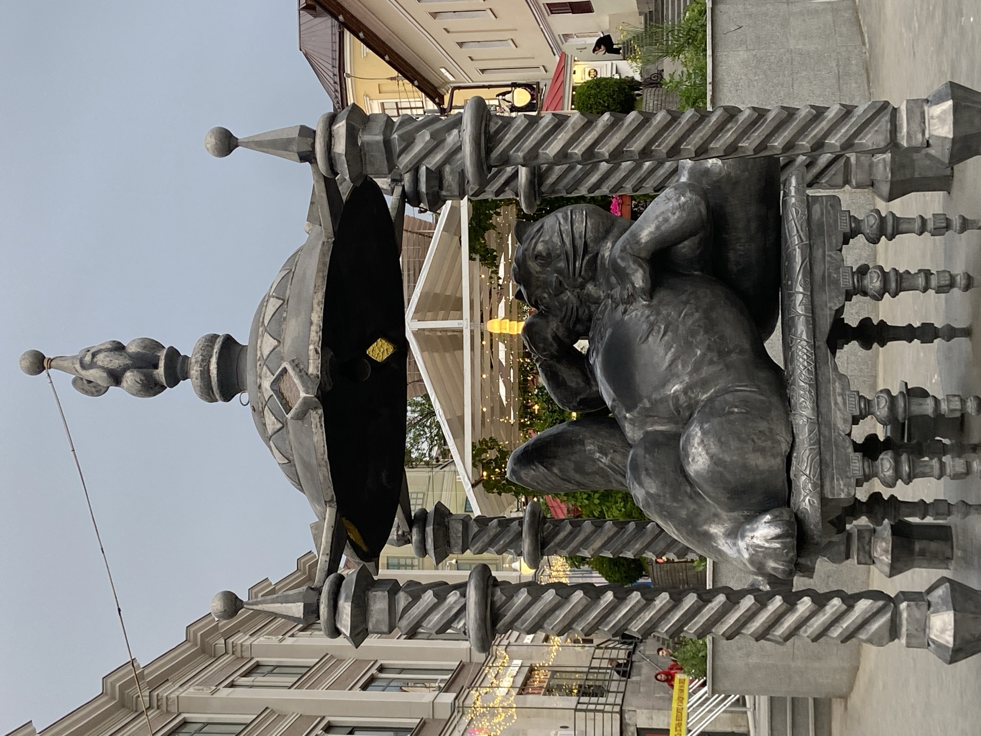 The image depicts a metal sculpture of a seated, draped figure, possibly allegorical, within an ornate, gazebo-like metal structure with a dome-shaped top. The sculpture is located outdoors, with commercial buildings and street lights visible in the background. The image is taken at an angle, the surroundings indicating an urban setting, likely Kazan, Russia. The setting appears to be a pedestrian area in the city.