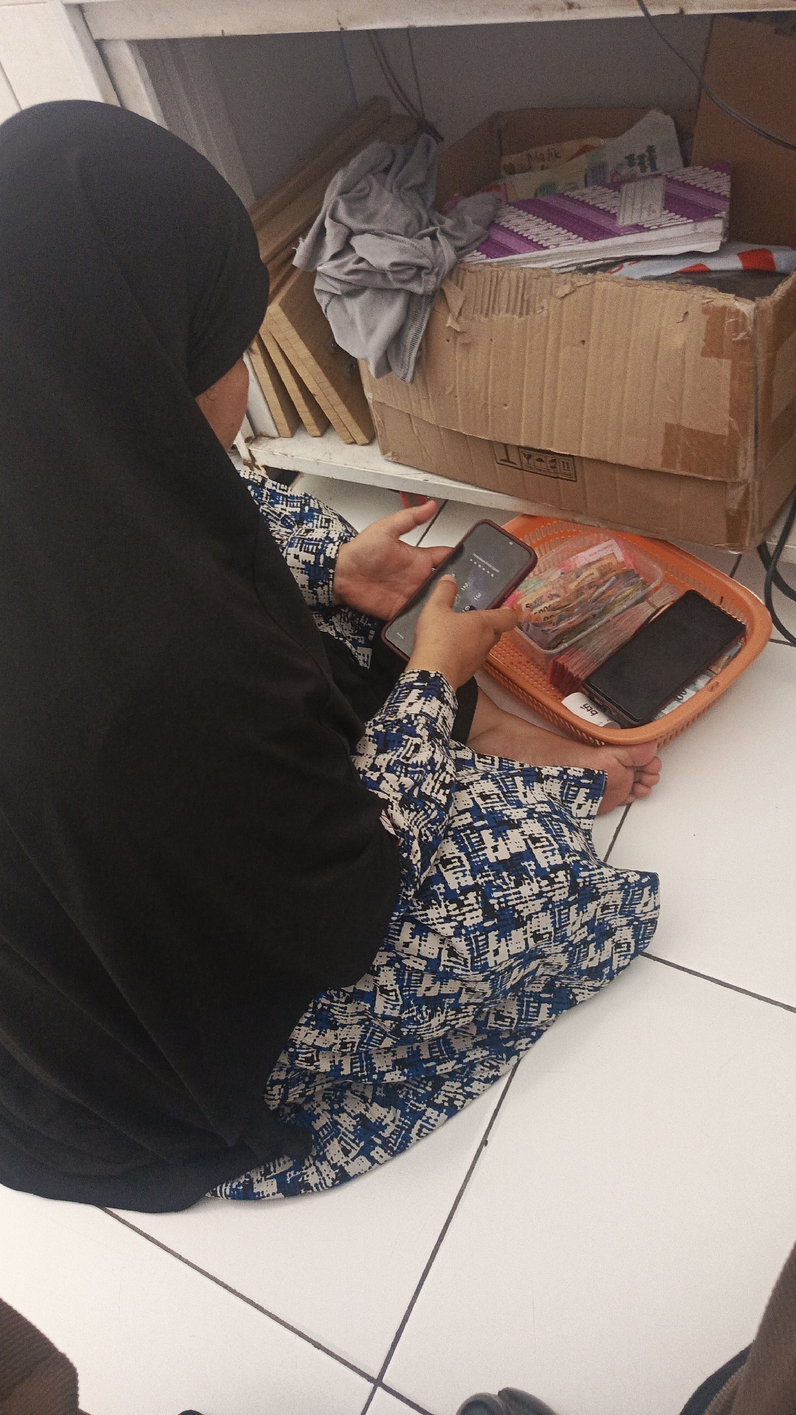 A person wearing a black hijab and a blue and white patterned dress is sitting on a tiled floor. They are holding a smartphone in their left hand, with their right hand reaching out to touch the screen. In front of them, on the floor, is an orange plastic basket containing several stacks of Indonesian Rupiah banknotes and another black smartphone. Behind the person, on a shelf, are wooden planks, a folded grey garment, and a cardboard box filled with various items, including notebooks and what appears to be more paperwork. The scene is indoors, with a white ceiling and tiled floor visible. The general environment suggests a storage or work area. The location is specified as Parung Panjang, Indonesia, though no specific visual cues within the image confirm this.