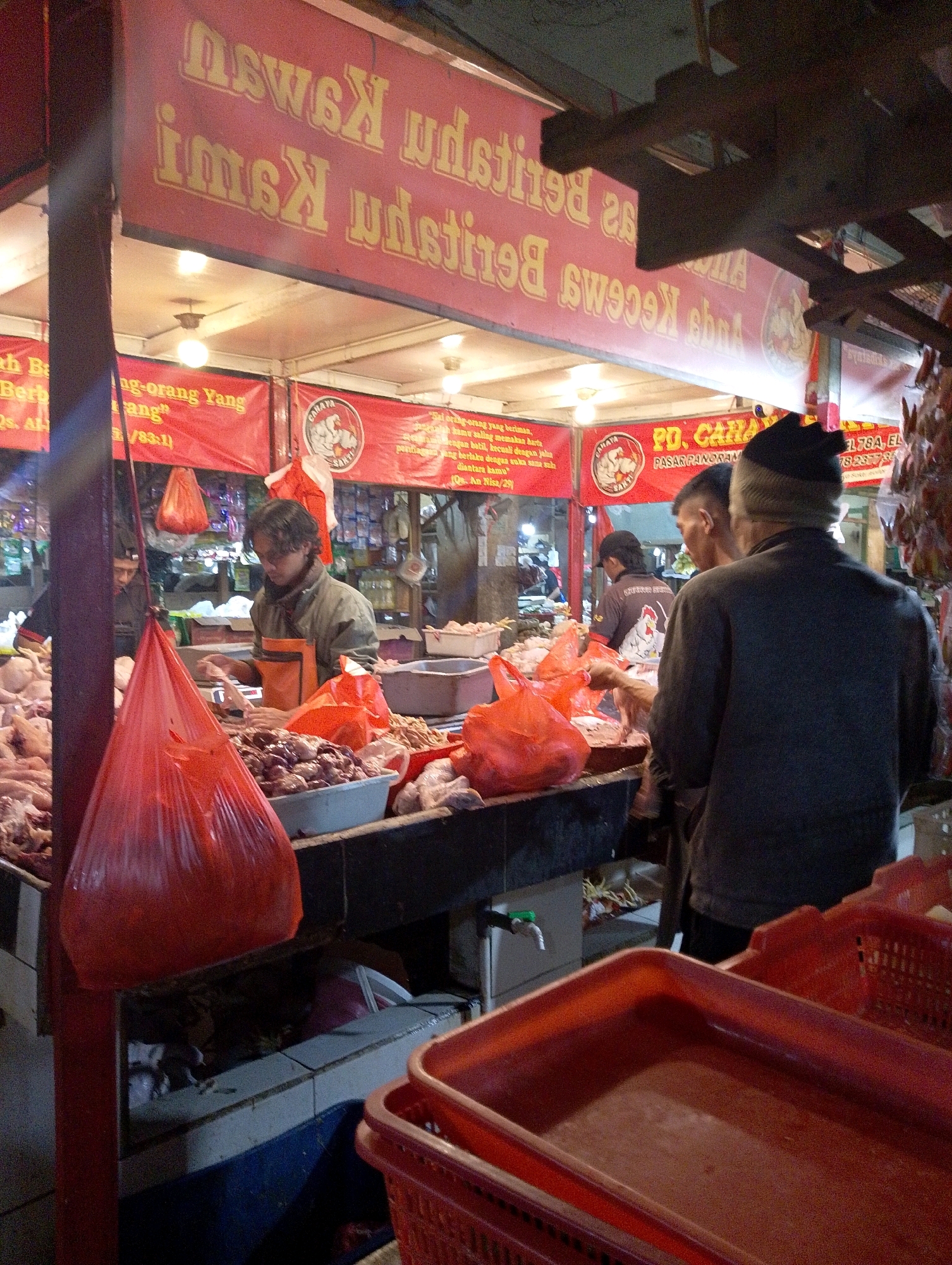 The image depicts a bustling outdoor market scene, likely in Lembang, Indonesia, given the signage and the types of goods displayed. In the foreground, a stall is laden with raw meats, primarily chickens and what appear to be offal items like livers, meticulously arranged in plastic containers and baskets. Several individuals are present, engaged in their respective activities. A man in an orange apron, presumably a vendor, is actively preparing or displaying meat. Another person, wearing a dark jacket and a beanie, is facing away from the camera, seemingly interacting with the vendor. In the background, more stalls with various food items and supplies are visible, illuminated by artificial lighting, suggesting the market operates into the evening. Red banners with yellow text are prominently displayed, likely advertising products or business names. The overall impression is one of a vibrant, working market environment.