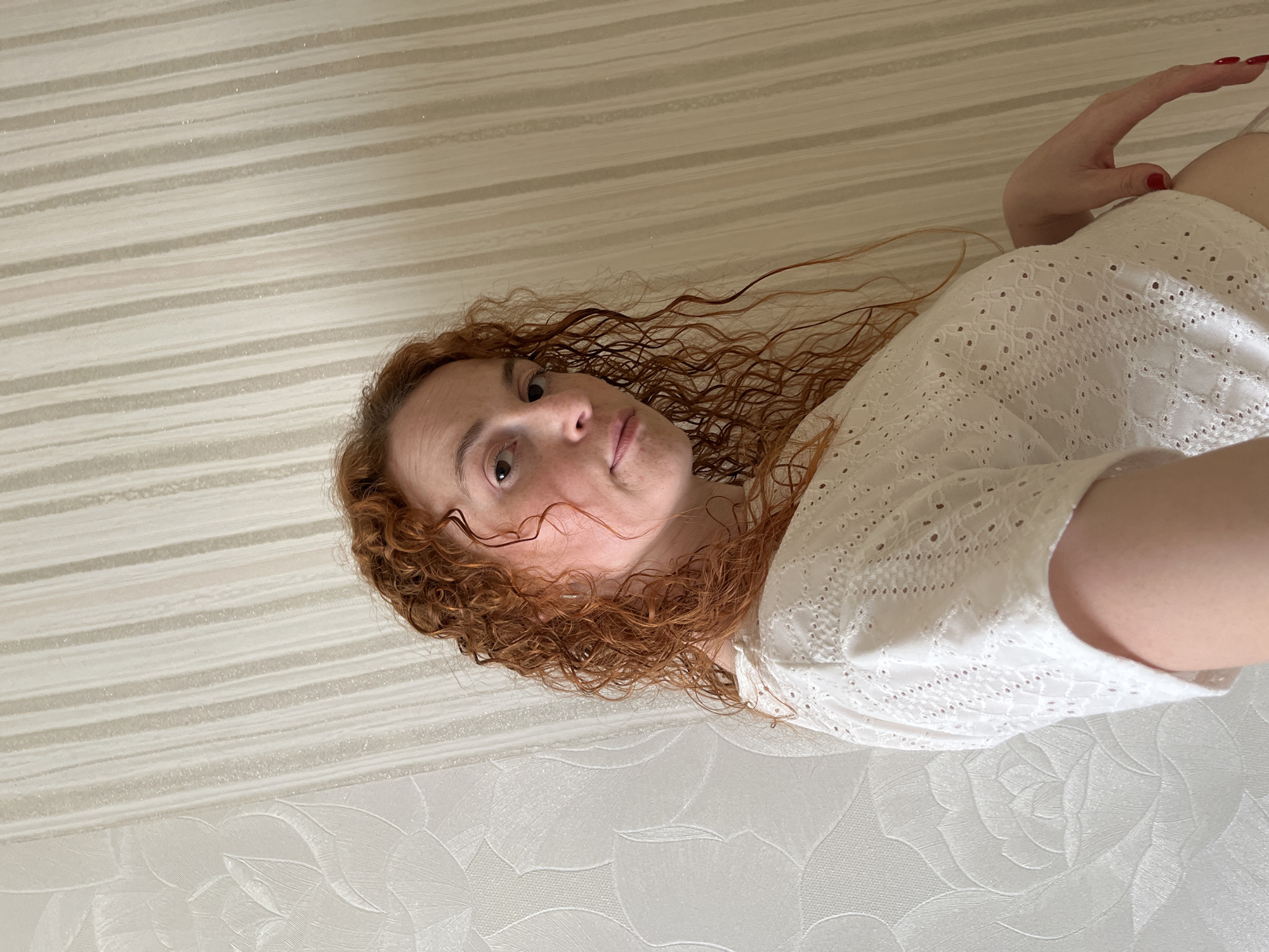 A person with red curly hair, wearing a white eyelet top, is seen from a low angle. The background is a vertically patterned wall, with a floral pattern at the bottom. A hand with red nail polish is visible in the top right corner. The person is looking towards the camera with a neutral expression. It is not possible to verify the location as Chaikovsky, Russia, from the image alone.