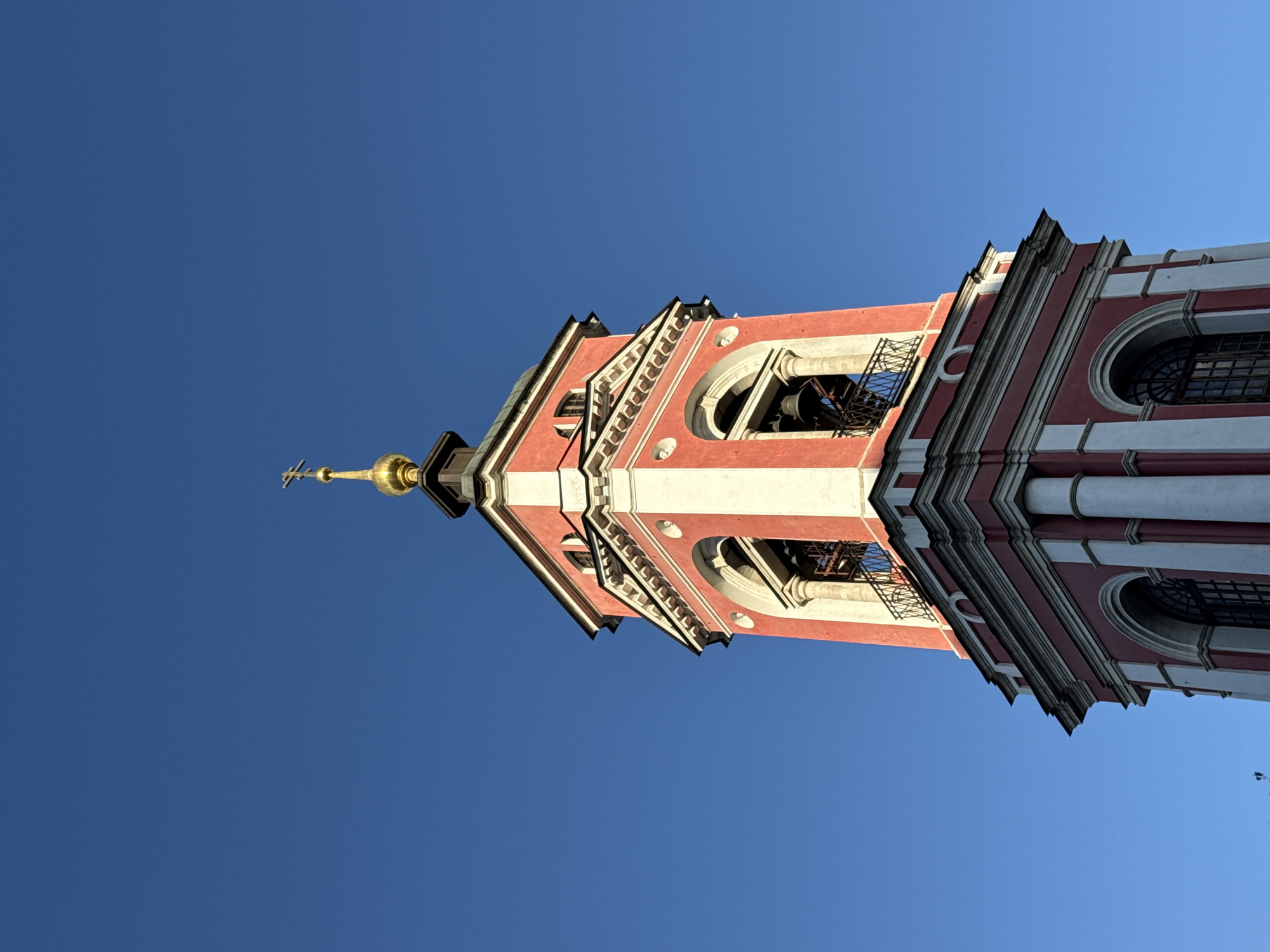 The image is a low-angle shot of the top of a bell tower, which is part of a church or cathedral. The tower is painted red and white, with decorative architectural details such as arches, columns, and intricate cornices. Bells are visible through arched openings, and a golden spire with a cross crowns the structure. The sky is a clear, vibrant blue, suggesting a sunny day. The image is oriented vertically, as if looking up from the base of the tower. The light and shadows indicate that the sun is high in the sky, likely midday. There are no people or animals visible. The overall impression is one of grandeur and religious architecture against a bright, expansive sky. The location is identified as Moscow, Russia.