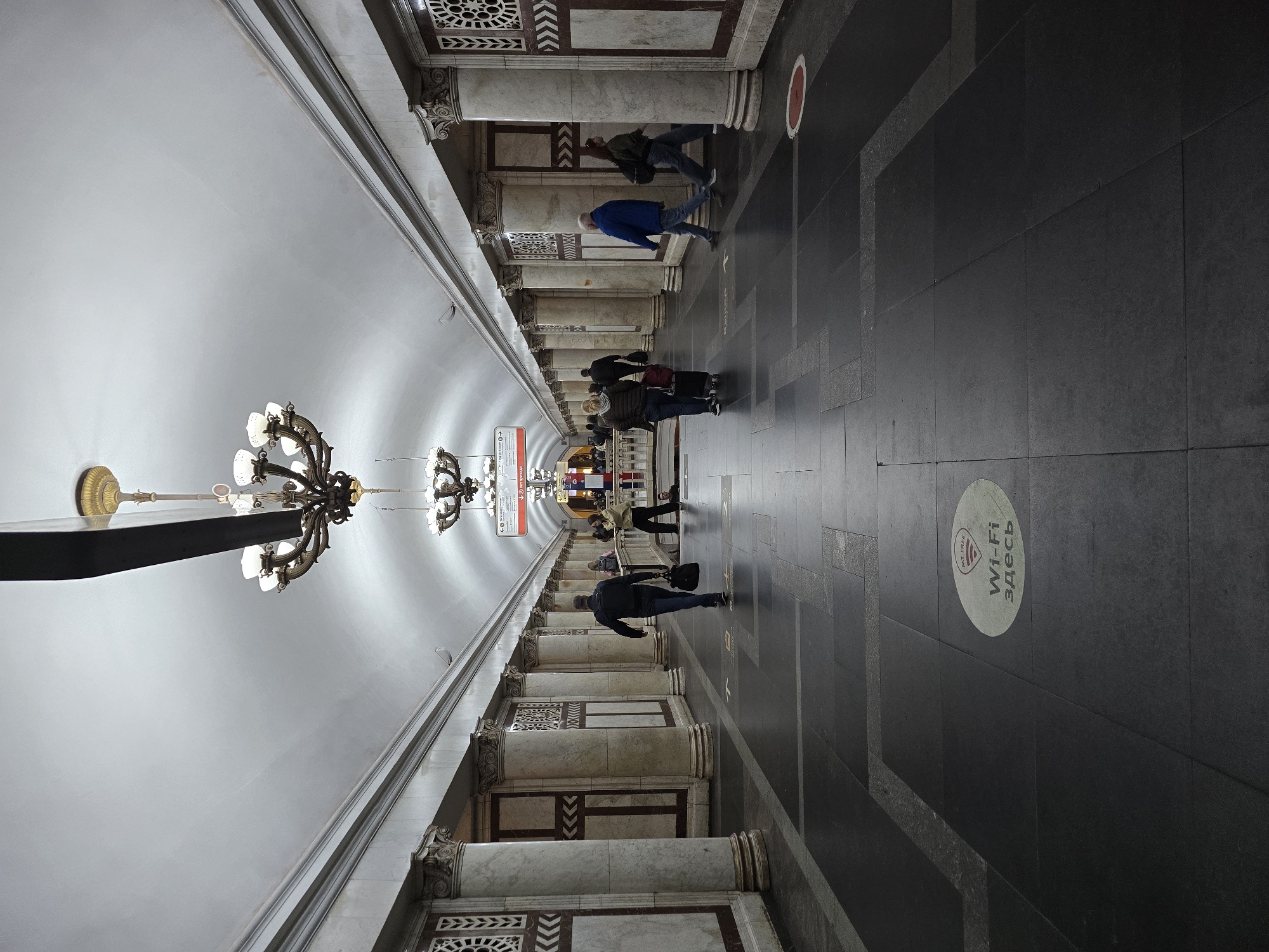 This is an interior shot of a Moscow Metro station in Russia, specifically the Kiyevskaya station on the Arbatsko-Pokrovskaya Line. The perspective is lengthwise down the center of the platform. The scene is brightly lit. The ceiling is arched and white, with hanging chandeliers. On either side are columns with decorative panels. People are walking along the platform, some with luggage. There are circular markers on the floor. A sign indicates "Wi-Fi здесь" ("Wi-Fi here" in Russian).