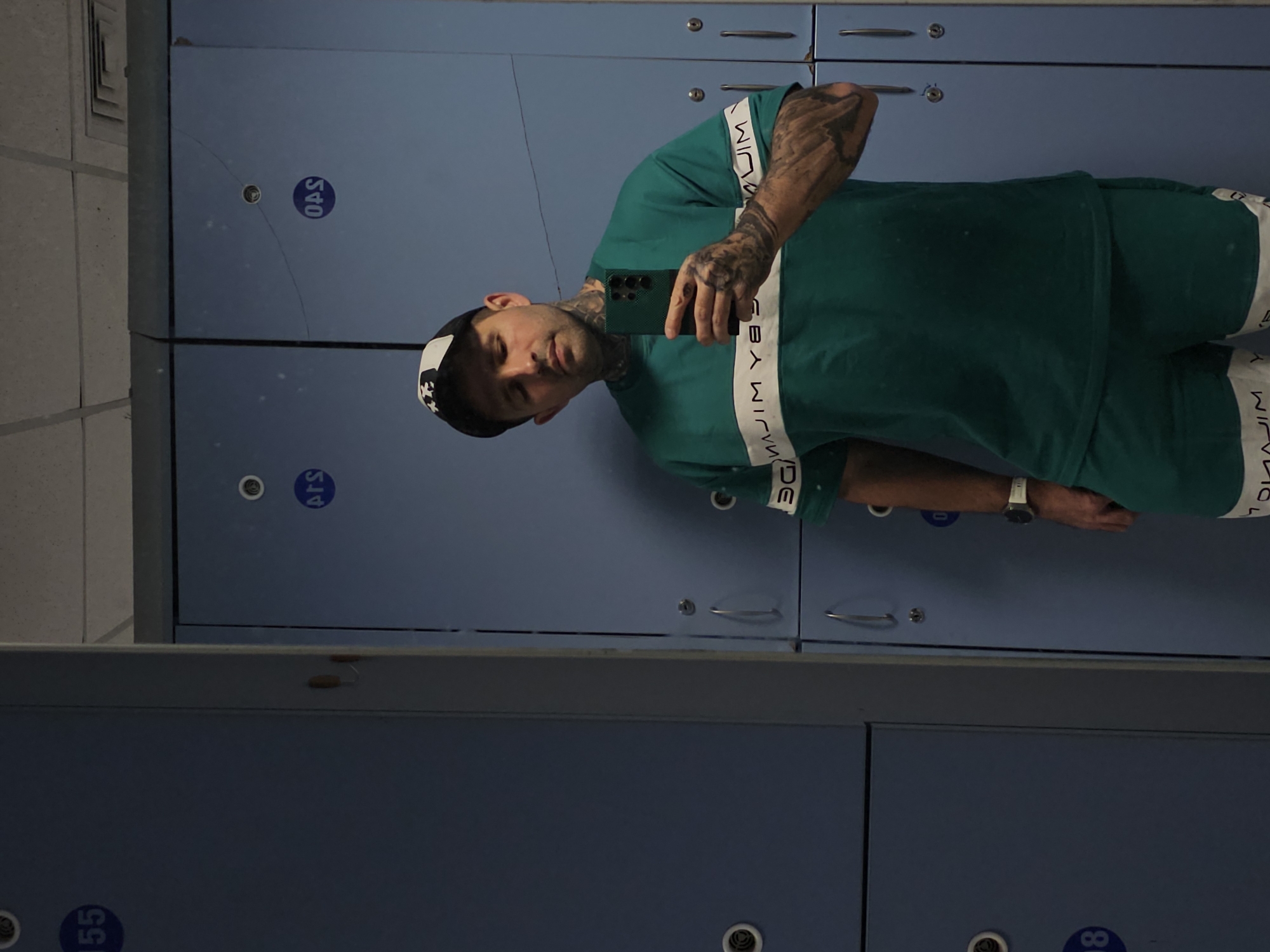 In a locker room, a man takes a selfie in front of blue lockers. He is wearing a green t-shirt with white stripes and a baseball cap. The lockers are numbered, with one visible locker labeled "155" on the left side of the image. Another locker in the background is labeled "OAS." The man has tattoos on his neck and arms and is holding a smartphone in his right hand. The environment appears to be clean and well-lit.