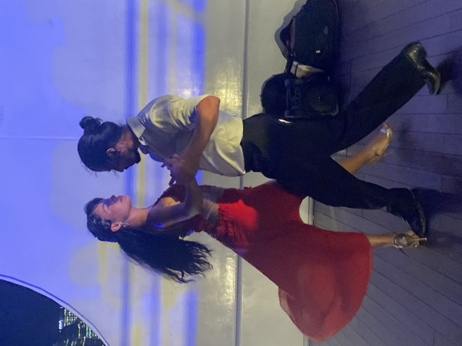 Two tango dancers, a female and a male, are captured mid-performance on a dark, reflective wooden floor, likely aboard a vessel in Puerto Madero, Buenos Aires, Argentina. The background reveals a curved white interior wall illuminated with horizontal blue and yellow strips, through which the distant city skyline, including an illuminated "BBVA" building, is visible under a night sky.
The female dancer, positioned on the left, wears a sleeveless, low-cut red gown with a flowing skirt that trails behind her left leg, which is extended backward. Her dark hair is styled partly up with visible loose curls, and she wears earrings and light-colored high heels. Her gaze is directed towards her partner.
The male dancer, positioned on the right, wears a light-colored collared shirt with sleeves rolled up and dark trousers with dark dress shoes. His dark hair is pulled back into a bun. He is also looking towards his partner.
The dancers are in a close embrace; the male's left arm is around the female's waist/lower back, and his right hand holds her left hand. Both exhibit a dynamic posture consistent with a tango step. To the right of the male dancer's feet, two dark bags or cases are present on the floor, one resembling a speaker.