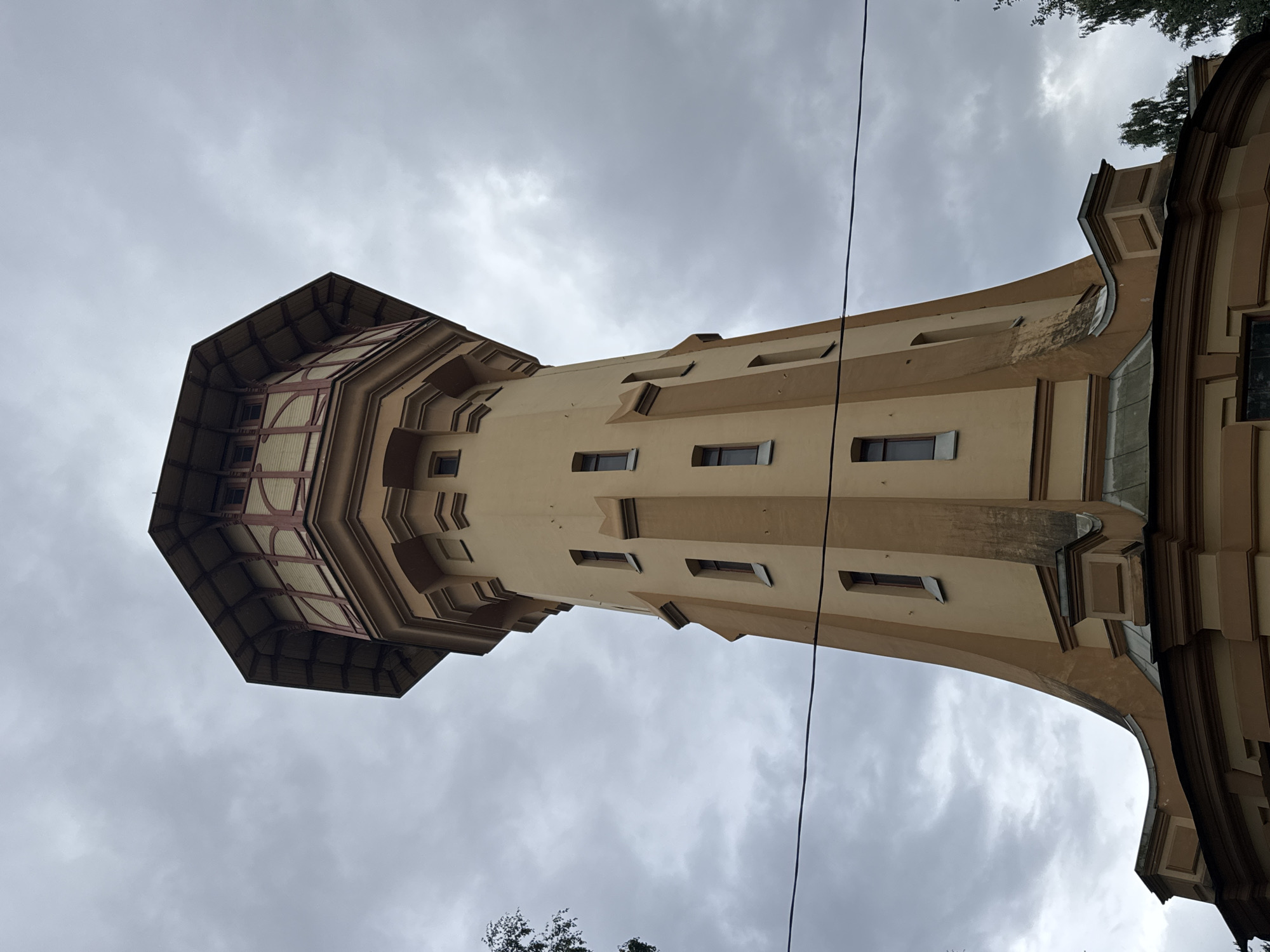 An eye-level, low-angle shot captures a tall, octagonal water tower made of light brown material against a cloudy sky. The tower features multiple windows, some narrow and vertical, others square. The uppermost section of the tower is capped with a dark, sloped roof, supported by decorative wooden beams and panels. The base of the tower is wider and circular, with a multi-layered roof structure. A dark wire runs horizontally across the frame in the lower half of the image. Trees are visible on the right side of the frame, partially obscuring the lower portion of the tower. The scene appears to be in an outdoor urban setting.