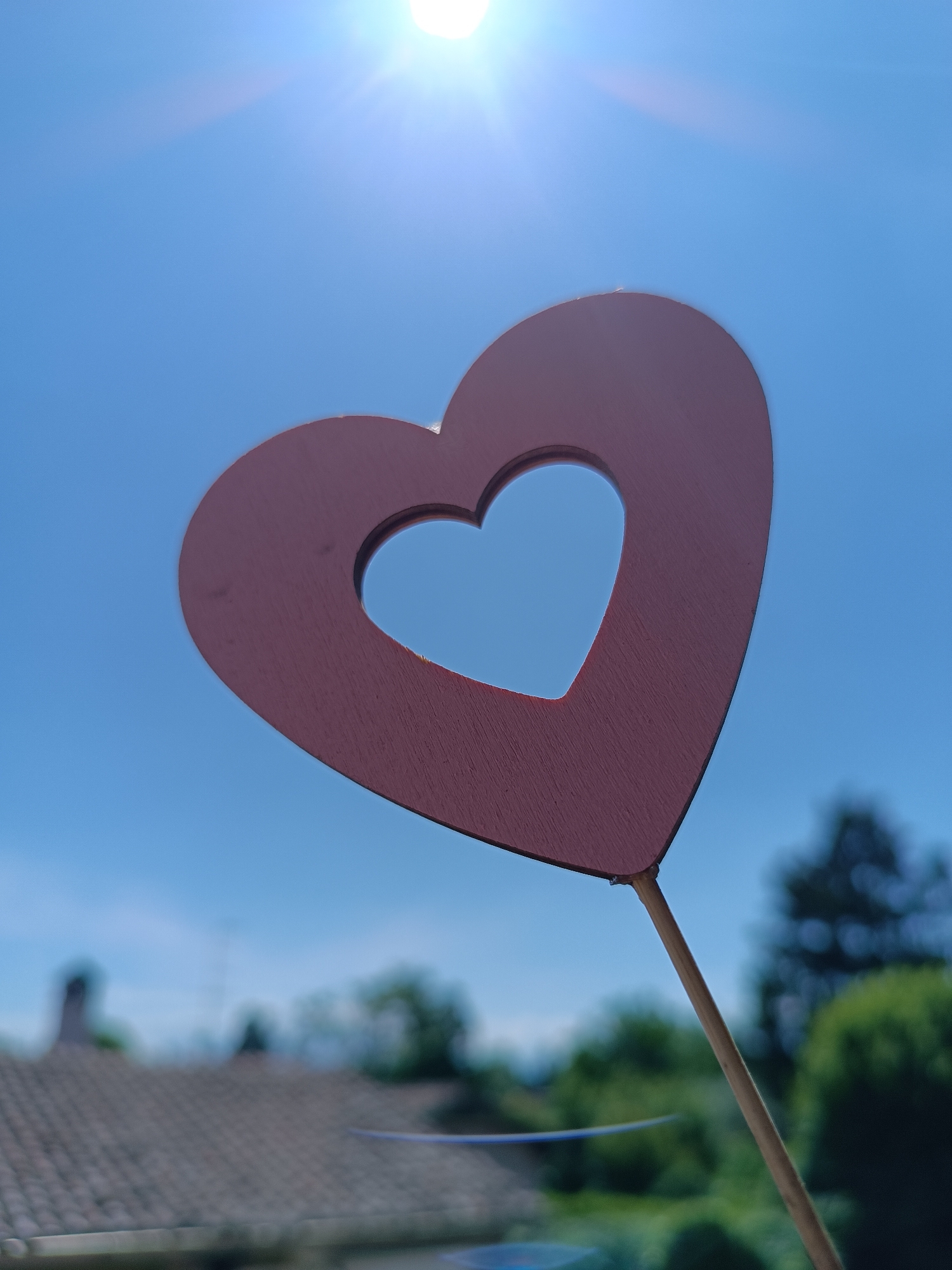 The image depicts a wooden heart-shaped cutout on a stick, held against a bright blue sky with the sun visible at the top. The heart is reddish-brown in color and has a smaller heart-shaped cutout in its center. The stick appears to be made of wood. The background includes a blurred view of rooftops and greenery, suggesting a residential area. Based on the prompt, this is likely a scene in Viadana, Italy, although specific identifying features confirming the exact location are not discernible from the image. The image is taken during the daytime, indicated by the bright sunlight and clear sky. There are no people visible in the frame.