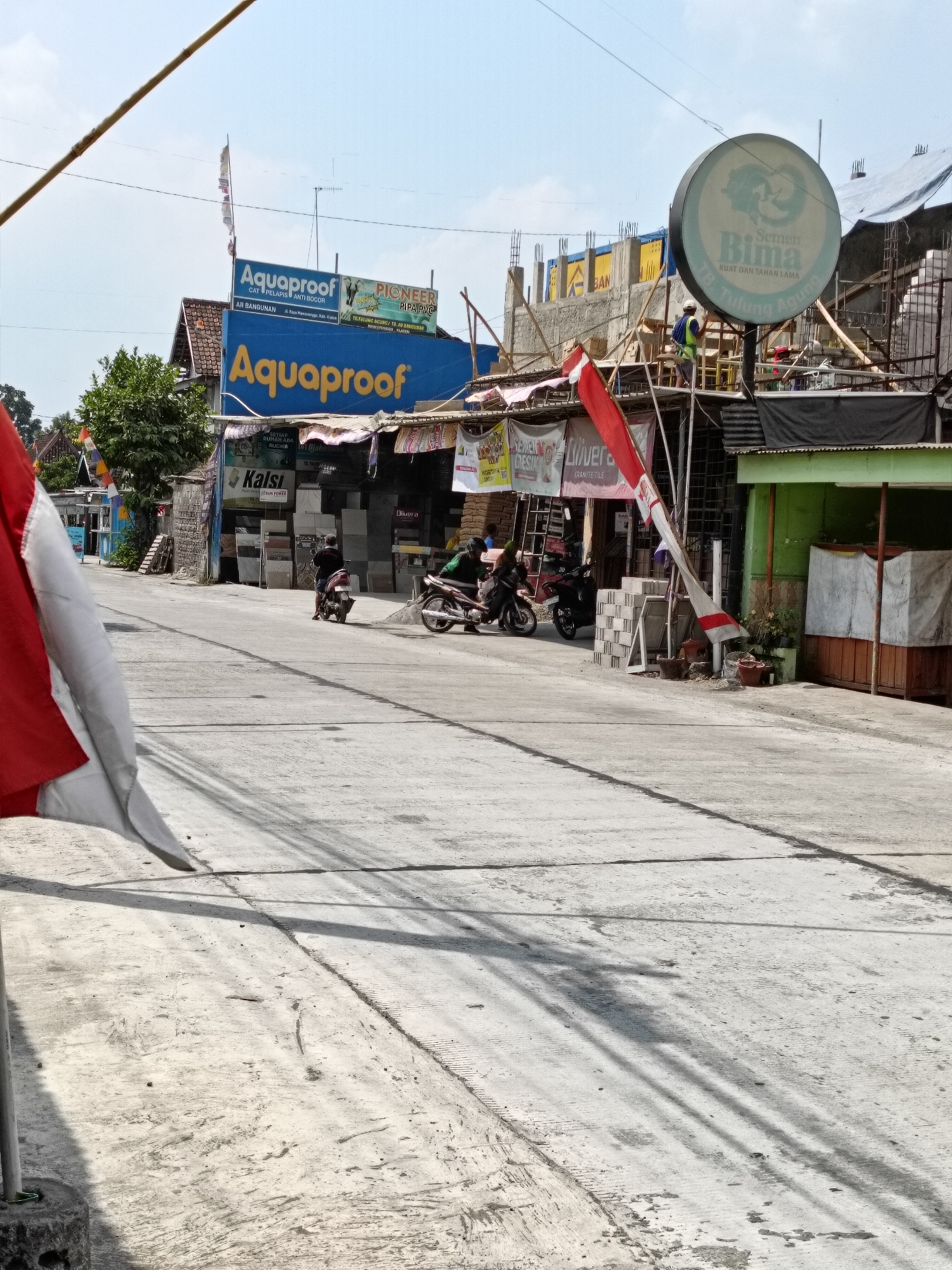 In Manisrenggo, Indonesia, a street scene unfolds under a clear sky. The road, paved with concrete, is partially shaded by a flag and overhead wires. On the right side of the road, a construction site is visible, with a worker in a green vest and hard hat in the process of building. A large, circular sign with the text "Bima Semen Kuat Dan Tahan Lama" hangs above the construction site. Adjacent to the construction, a store displays various tiles and building materials, with the brand "Aquaproof" prominently featured on its facade. Two motorcycles are parked on the side of the road, and two individuals are riding motorcycles, one with a passenger, moving along the street. The overall environment suggests an active commercial and construction area within the city.