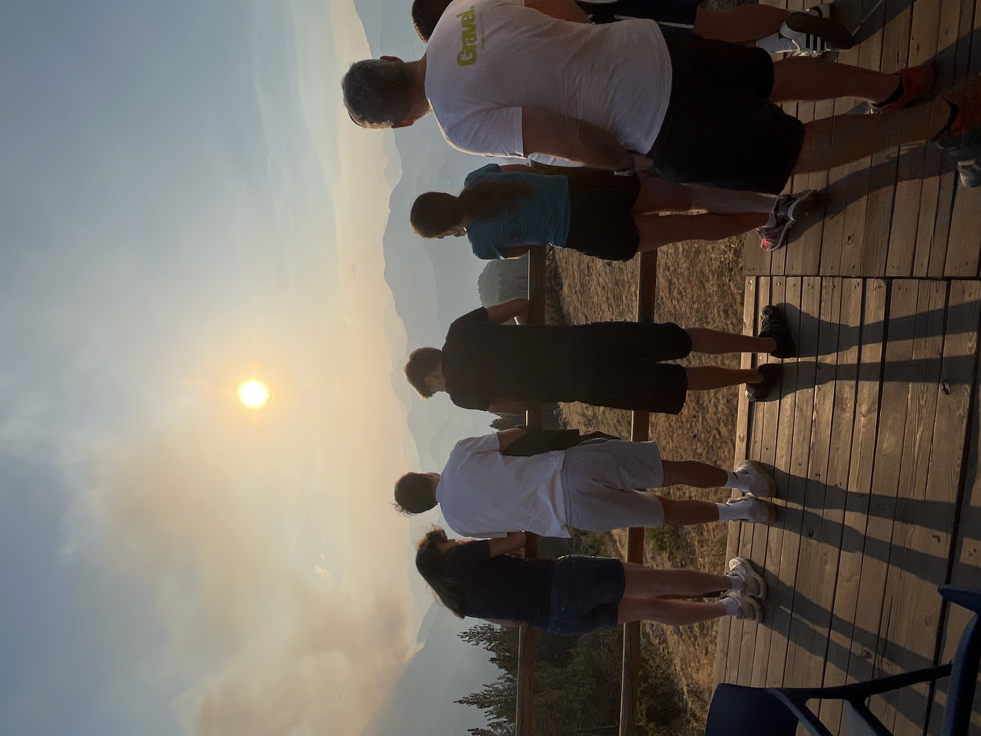 The image shows a group of six people standing on a wooden deck, looking out at a mountainous landscape. The sun is visible in the sky, partially obscured by smoke, suggesting a possible wildfire in the distance. The people are casually dressed in t-shirts and shorts. The person on the far right is wearing a white t-shirt with green lettering that reads "Gravel". The location is identified as San Juan de Plan/San Chuan de Plan, Spain. The overall lighting suggests it is late afternoon or early evening, with long shadows cast across the wooden deck. The environment appears to be a natural, possibly elevated, viewpoint overlooking valleys and mountain ranges.