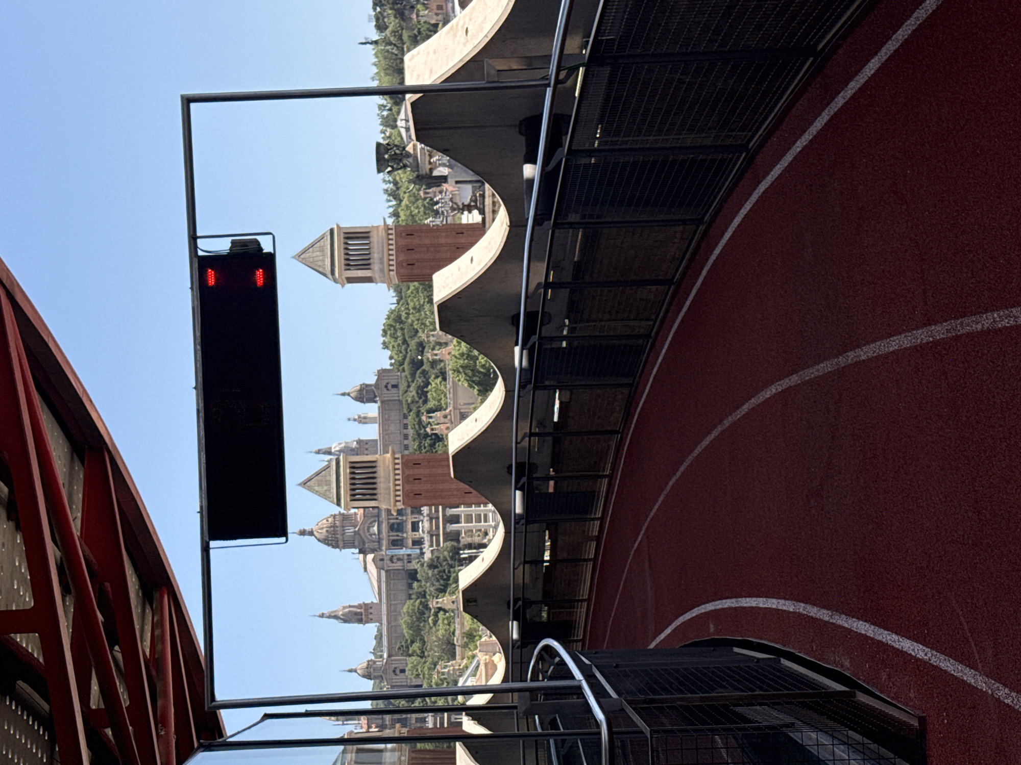The image shows a red running track with white lane lines curving to the left. A metal railing with a dark mesh fence runs along the outer edge of the track. Overhead, a red metal structure with visible beams is present. In the background, a cityscape is visible under a clear blue sky. Prominent architectural features include domed buildings and tall, tower-like structures with classical architectural elements. A black digital display with red lights is mounted on a metal pole in the upper right quadrant. The architectural style and the presence of structures reminiscent of the Palau Nacional suggest the location is near Montjuïc in Barcelona, Spain. There are no people or animals visible in the image.
